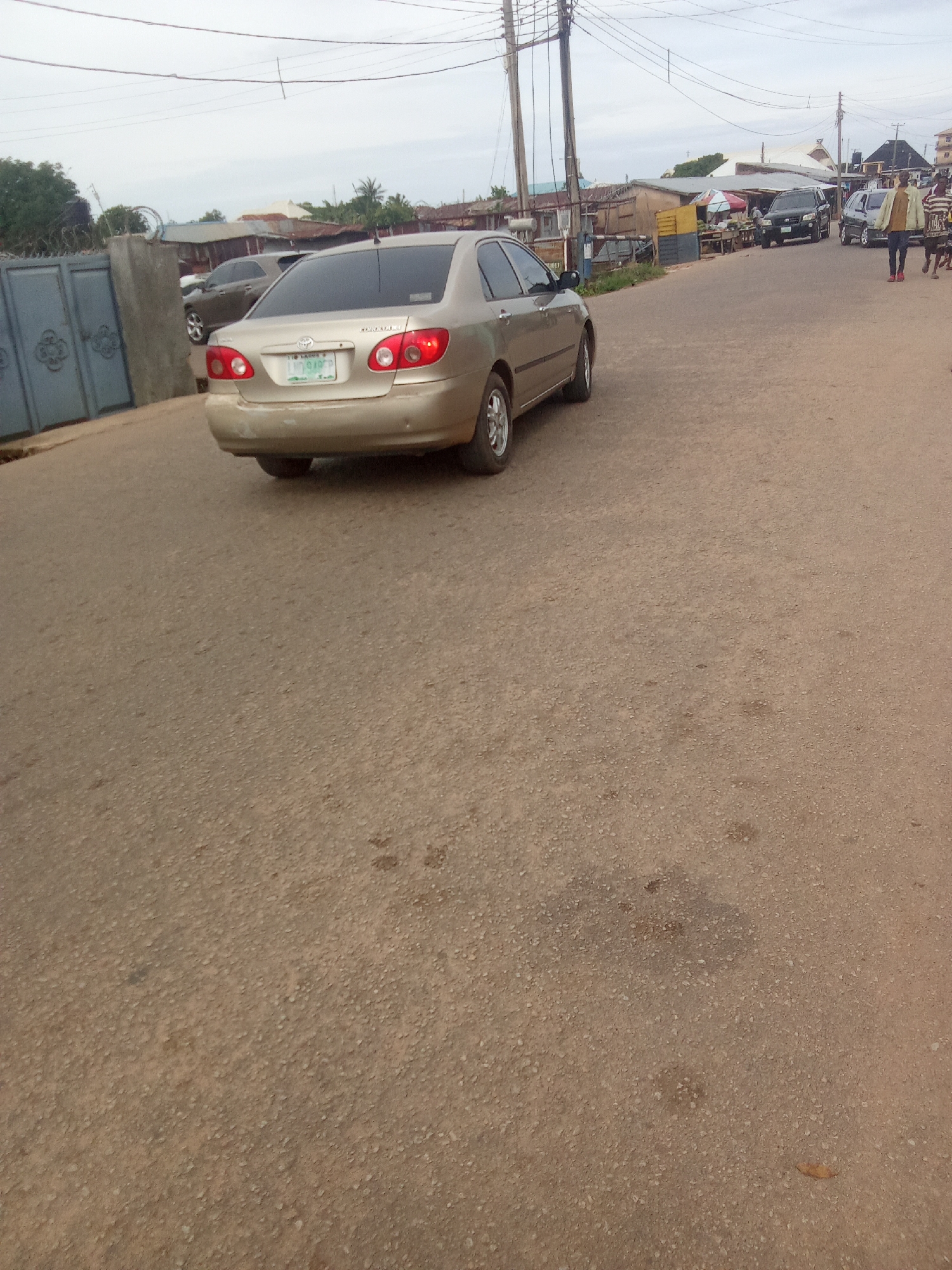 The image depicts a street scene in Jos, Nigeria, on an overcast day. A gold-colored Toyota Corolla is parked on the right side of the road, facing away from the camera. The car's license plate is visible, showing "LGT 949 PP". In the background, several other vehicles, including a black SUV and a white car, are present, along with buildings and market stalls. A person in a light-colored jacket and dark pants is walking on the sidewalk. The road surface is paved with asphalt and appears to be in moderate condition. The overall atmosphere suggests a typical urban environment in Nigeria.
