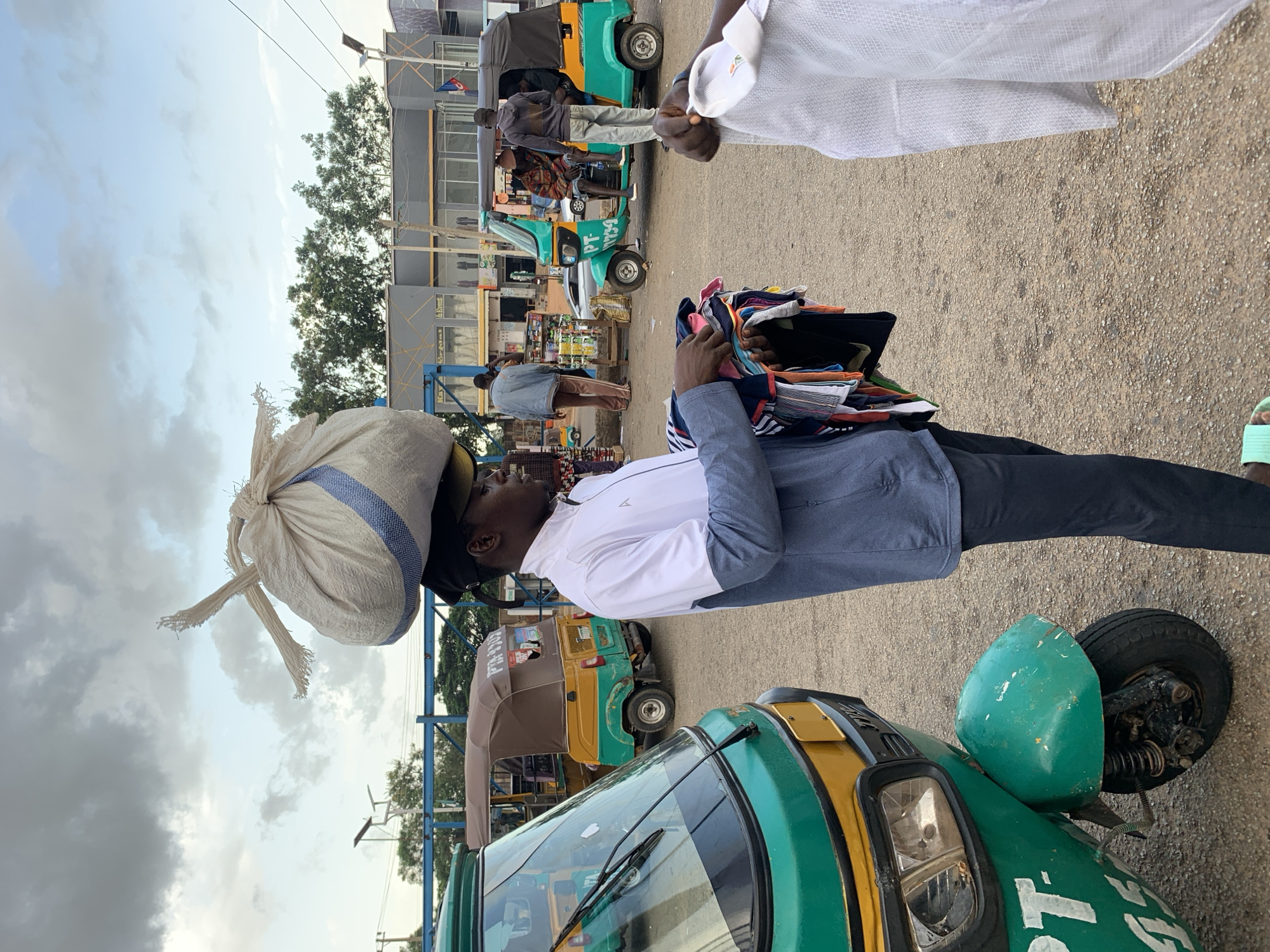 A man with a large sack balanced on his head walks in what appears to be a bustling market area in Jos, Nigeria. He is holding a bundle of folded clothing. The scene is set against a backdrop of a cloudy sky and a row of buildings. Several auto-rickshaws, known locally as "keke napep," are visible in the background, suggesting a busy transportation hub. One auto-rickshaw is a vibrant green and yellow, with the license plate "PT 9739." Another, in brown and green, is parked nearby. Pedestrians are scattered throughout the area, with some appearing to engage in commerce. The ground is a dusty, gravelly surface. The man's attire consists of a white and blue jacket and dark trousers. The overall impression is one of everyday life and commerce in an urban Nigerian setting.