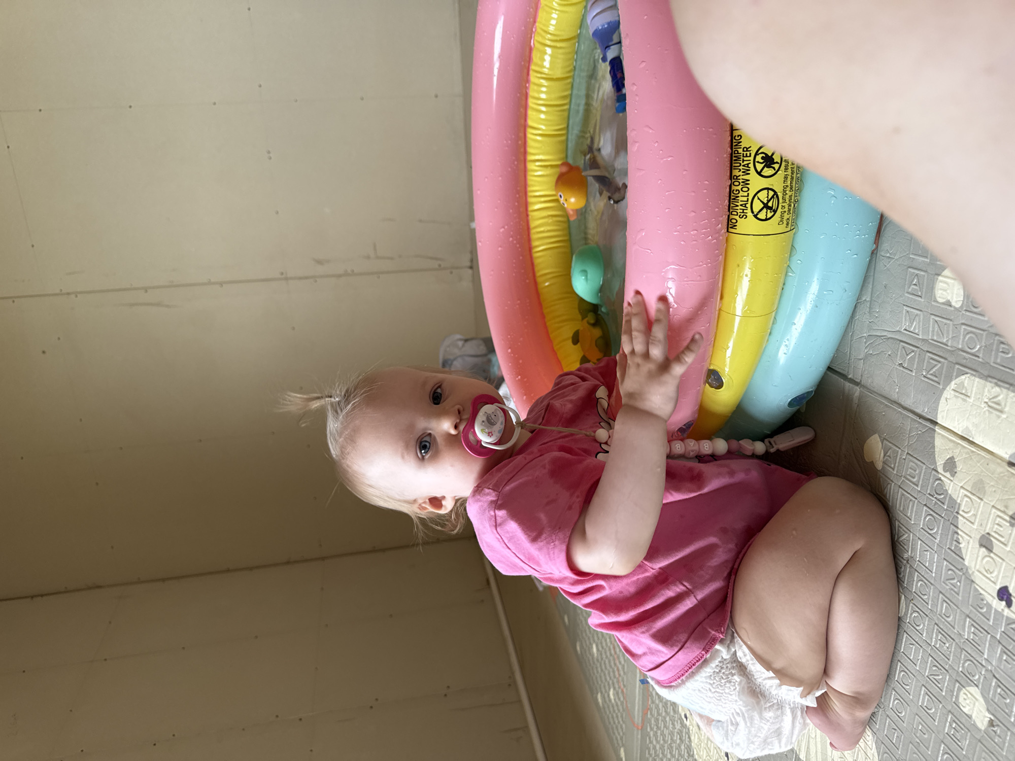 A baby is sitting on an alphabet-patterned floor mat next to a colorful inflatable pool. The pool is partially filled with water and toys. The baby, wearing a pink shirt, a diaper, and a pacifier, has blonde hair in a small ponytail. A fair-skinned arm is partially visible in the upper right corner. The location appears to be indoors against a wall. There is no indication of this being in the city of Фоки, Russia.