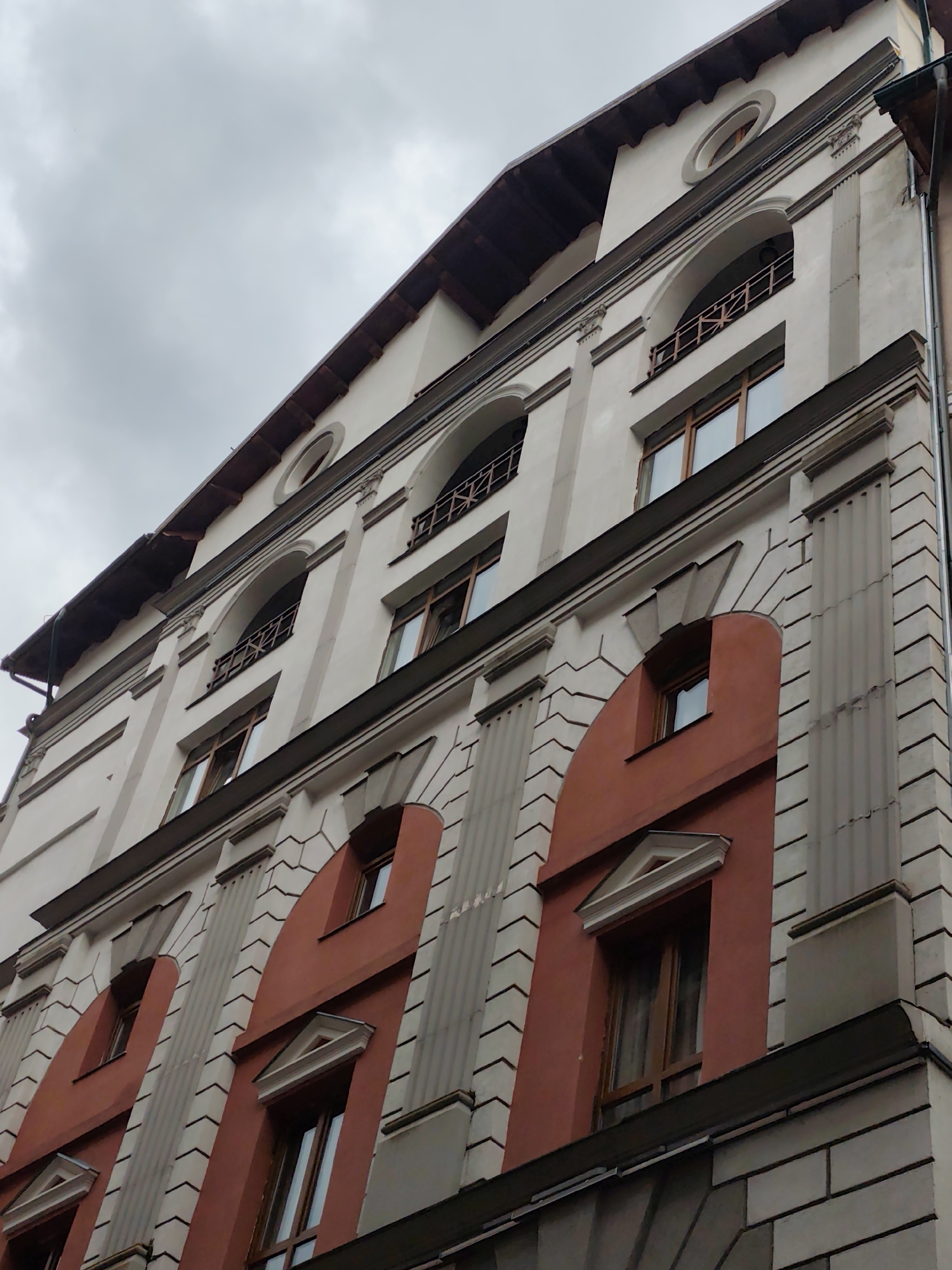The image shows a low-angle, full-shot view of the façade of a light-colored building with ornate architectural details, possibly located in Estosadok, Russia. The sky is overcast and gray, suggesting a cloudy day. The building has multiple stories, with several windows visible on each level.
The architectural style appears to be classical or neo-classical, with elements such as pilasters, arched window frames, and decorative moldings. Some sections of the façade are painted a deep rust or reddish-orange color, contrasting with the lighter main color. There are visible horizontal lines and patterns on the façade, creating a textured appearance.
The roofline is visible at the top of the building, featuring a dark wooden overhang supported by decorative brackets. The windows are rectangular, with wooden frames and dark panes. Some windows are set within arched recesses, and one upper-level window is circular. A balcony with a decorative metal railing is visible on one of the upper floors.
The image is a static architectural study, with no people or activity depicted. The focus is entirely on the building's exterior and its design features.