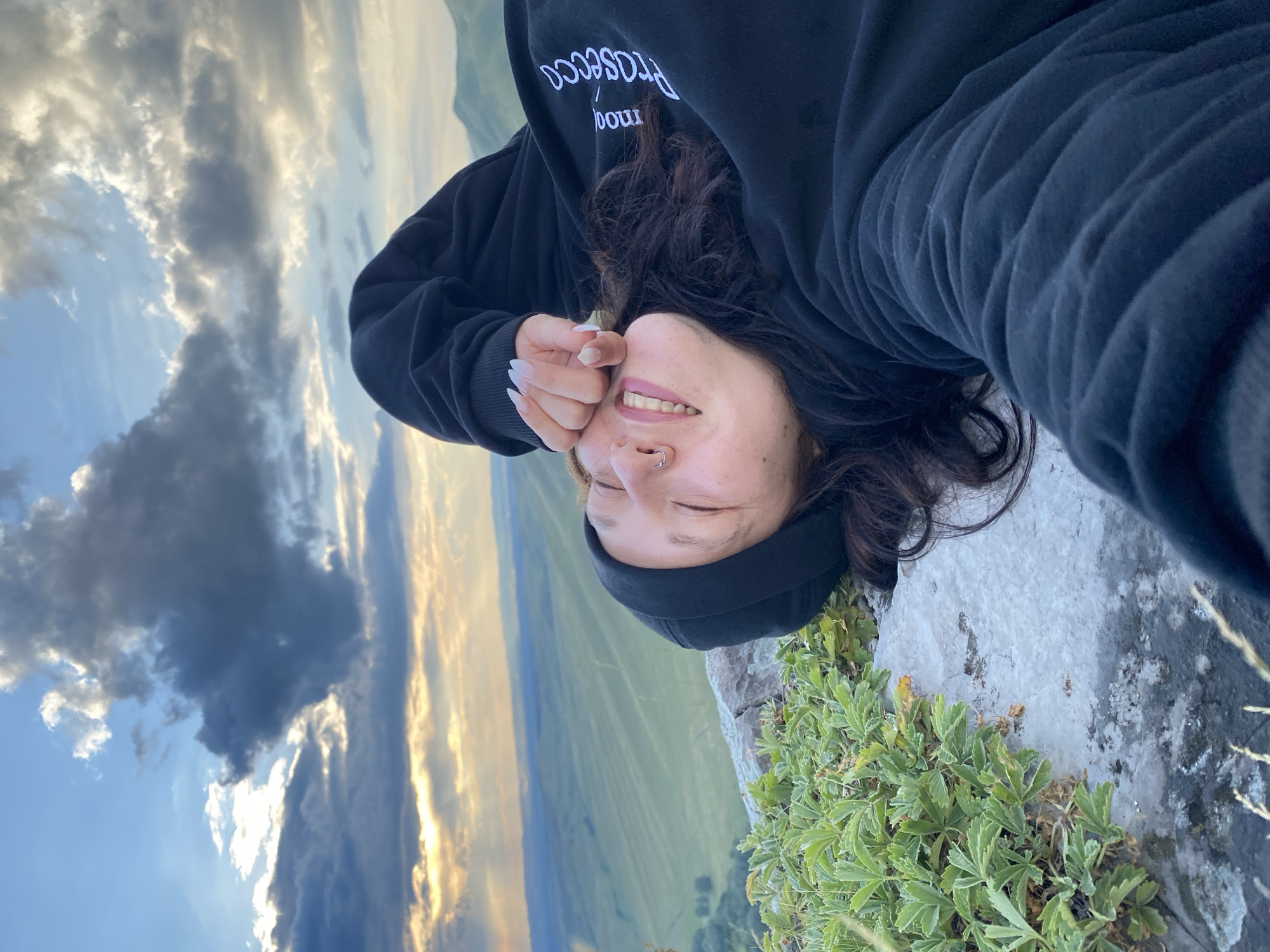 A young woman lies on her back on a rocky surface with green vegetation growing on it, facing the camera. She is wearing a black beanie and a black hooded sweatshirt. Her eyes are closed, and she has a smile on her face, with her right hand covering her forehead. She has a nose ring. The background consists of a vast, rolling green landscape under a cloudy sky with hints of sunset light filtering through. The scene appears to be in a natural, elevated location, possibly a mountain or high plateau. The location is identified as Abzanovo, Russia.