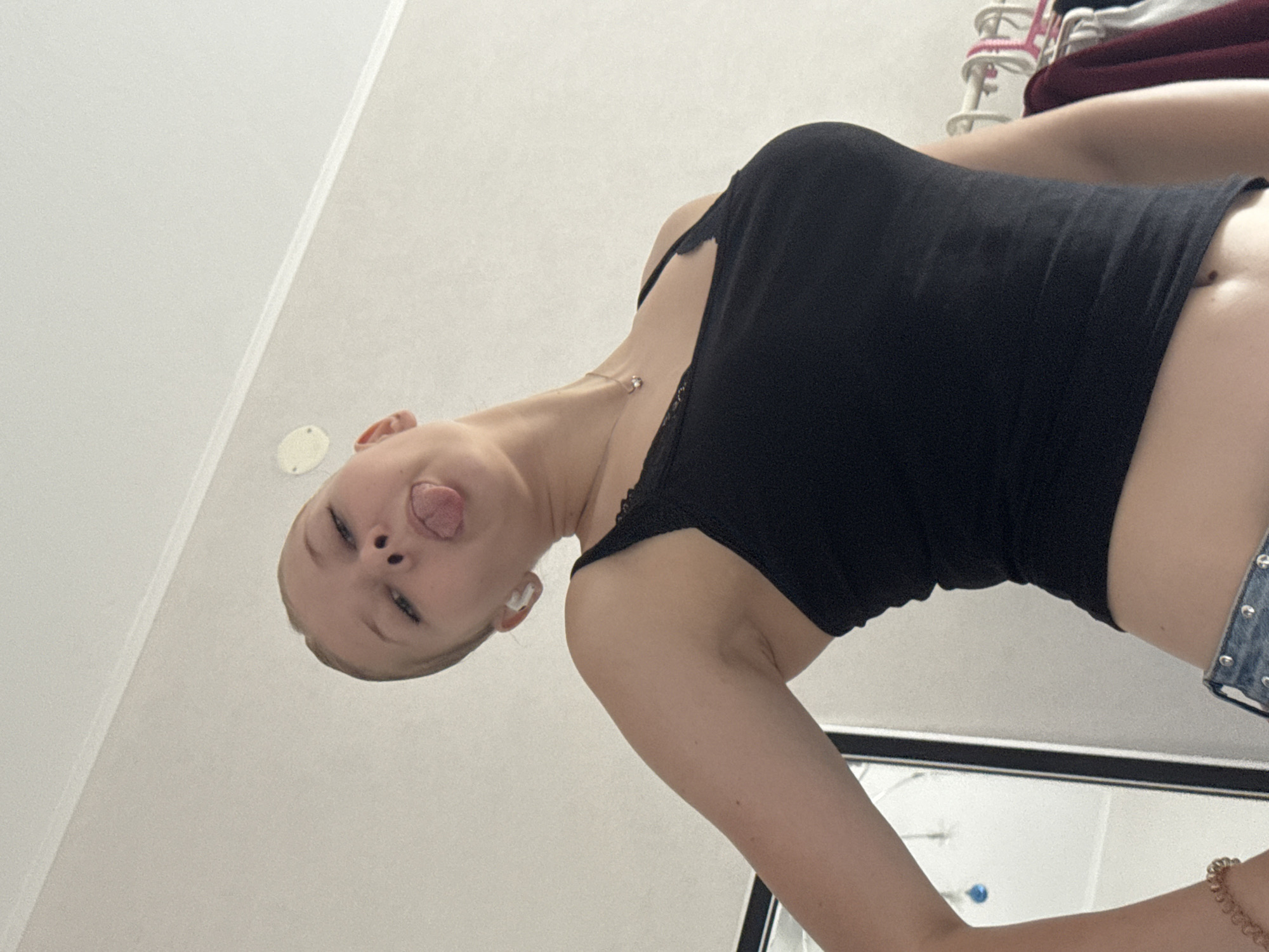 The image depicts a young person with blonde hair pulled back, sticking out their tongue and looking upwards. They are wearing a black tank top with lace trim and blue jeans. A thin necklace with a small pendant is visible around their neck, and a white wireless earbud is in their ear. The background shows a plain wall and a closet with clothes on hangers. The scene appears to be indoors, likely a bedroom, and there is no indication of the city of Perm, Russia.