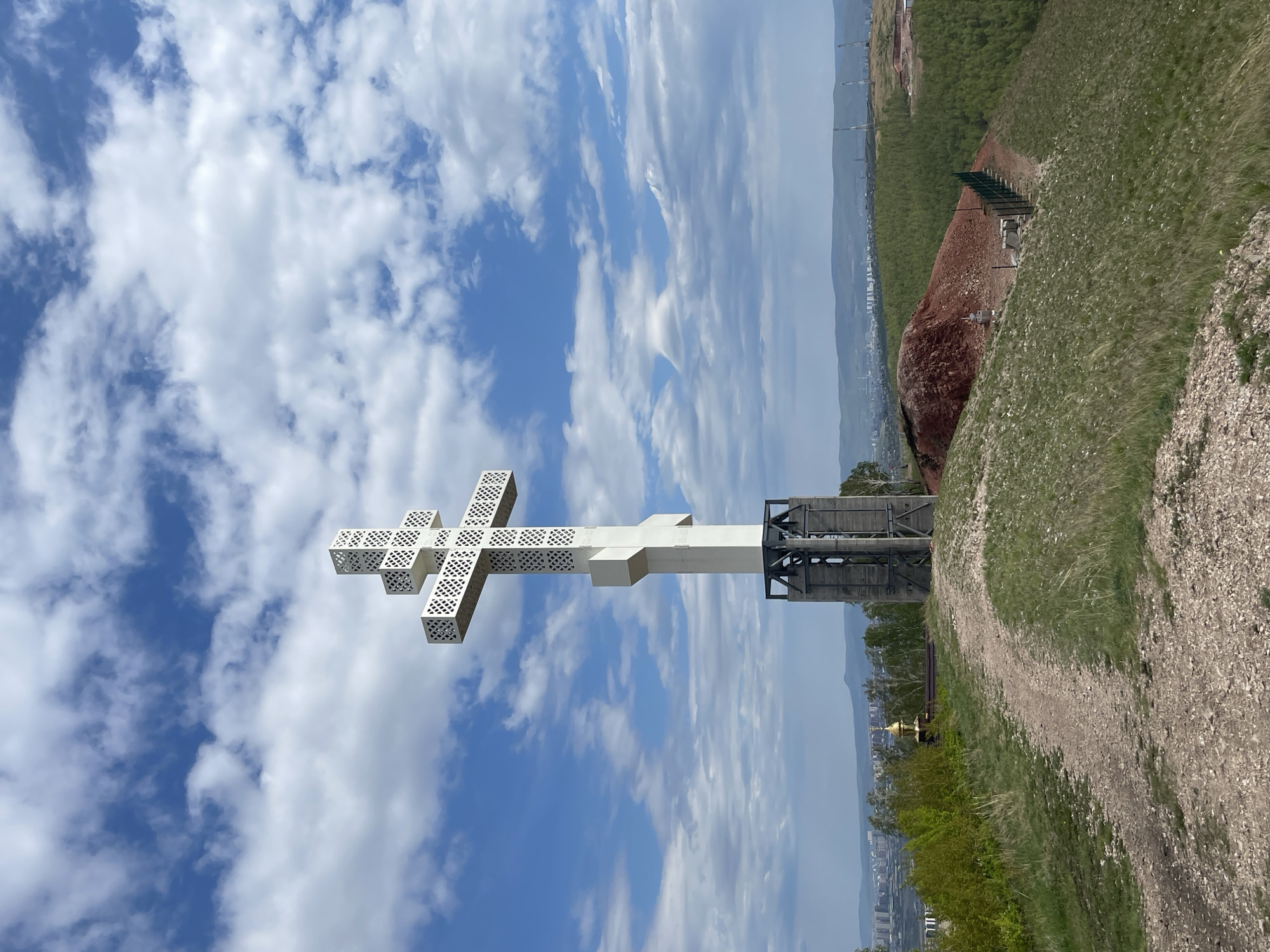 The image shows an Orthodox cross monument in Солонцы, Russia. The white cross, with a patterned design, is set against a partly cloudy blue sky. Below the cross is a metal structure that supports it. To the right of the structure, the landscape slopes downwards with grass and a rocky path. A small golden-domed structure is visible in the distance. The horizon shows the city of Солонцы.