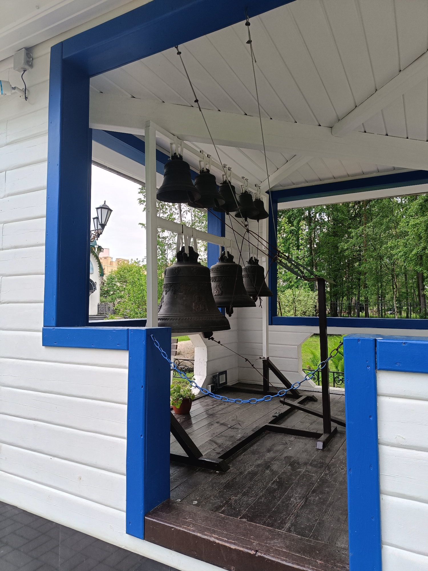 The image shows a series of bronze church bells hanging in a white and blue wooden structure. The structure has a wooden deck floor. The bells are arranged in a row, suspended by ropes. Outside the structure, green trees and greenery can be seen. In the background, a building is visible. The location is identifiable as the bell tower at the Alexander Nevsky Lavra in Saint Petersburg, Russia.