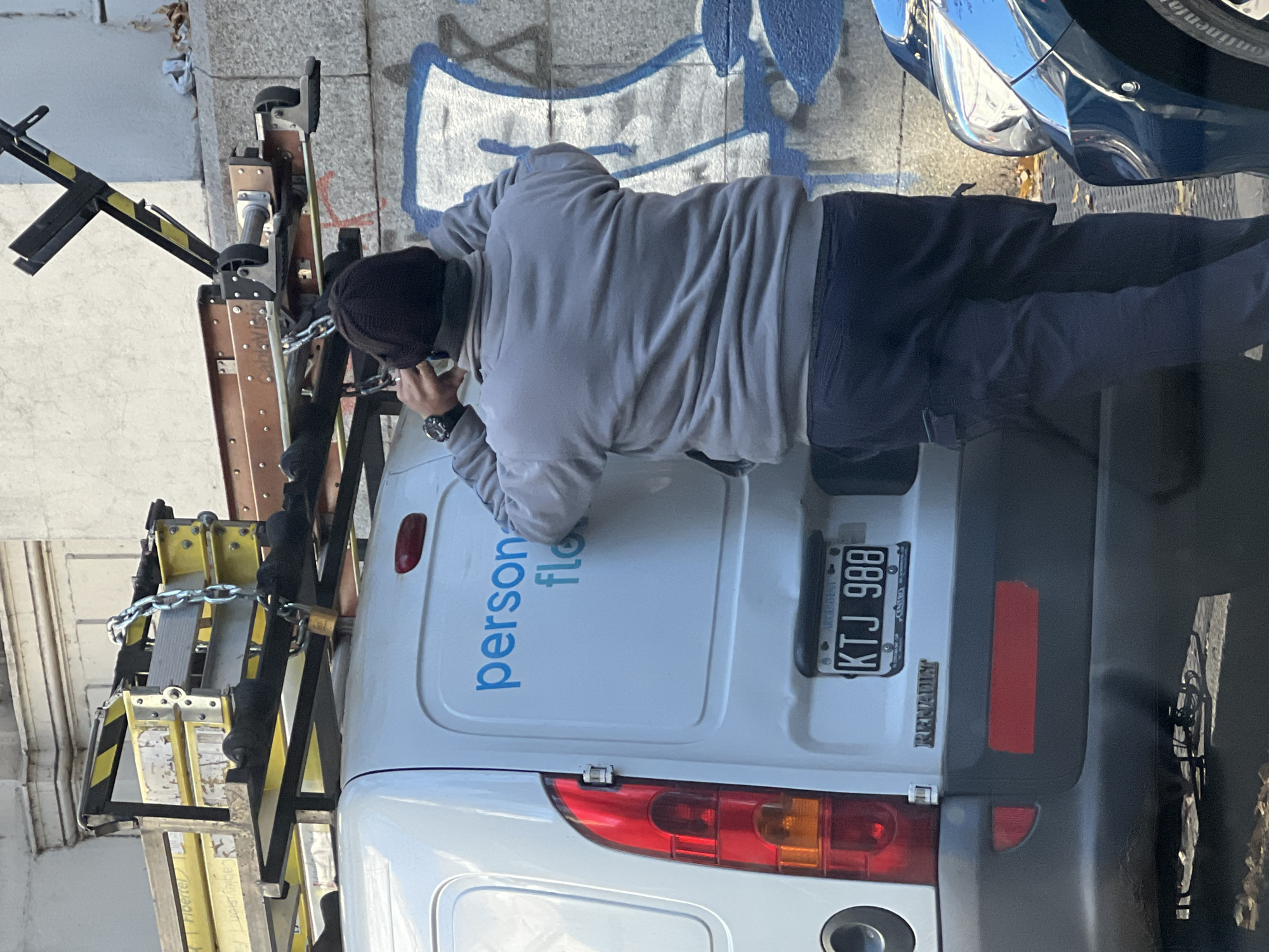 A man wearing a gray hooded sweatshirt and dark pants is looking at the rear door of a white Renault Kangoo van. The van has a ladder secured to the side with chains and a padlock. The van's rear door has the logo "personal flow" in blue lettering. The license plate reads "KTJ 988" and identifies the vehicle as being from the Autonomous City of Buenos Aires, Argentina. The background includes a building and a partial view of a blue car. Graffiti is visible on the building wall behind the van. The location appears to be an urban street in Buenos Aires, Argentina.