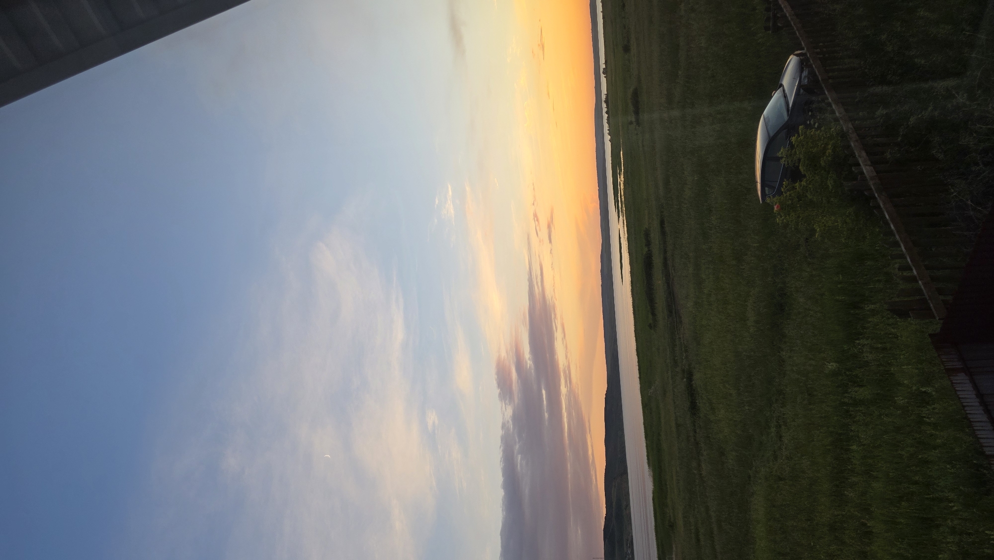 The image depicts a sunset over a wide body of water, likely a river or lake, with a silhouetted landscape on the opposite bank. The sky displays a gradient of colors, transitioning from a deep blue at the top to shades of orange and pink near the horizon, with wispy clouds scattered throughout. A sliver of a crescent moon is visible in the upper left quadrant of the sky. In the foreground, a grassy field stretches towards the water, with a wooden fence running along the bottom edge of the frame. A dark-colored car is parked in the field, partially obscured by tall grass and some foliage. The overall scene is tranquil and natural, suggesting a rural or undeveloped area. The location is identified as being in the city of Alga, Russia.