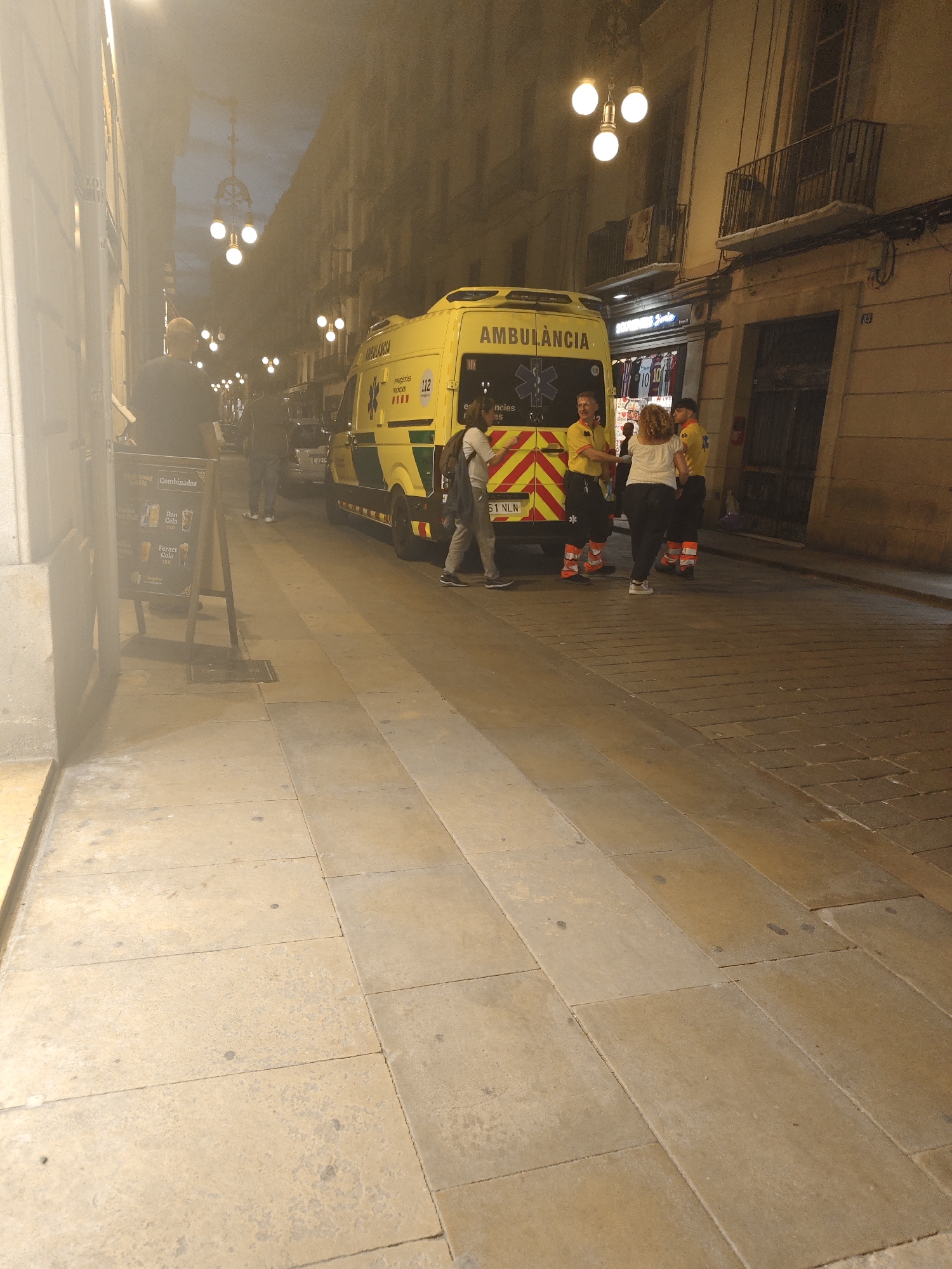 The image depicts a street scene at night in Barcelona, Spain, featuring an ambulance parked on the right side of the street. Four ambulance workers are gathered near the back of the vehicle, engaged in a discussion. A woman in a white top and dark pants is also part of the group. Other pedestrians are visible in the background, including a man standing near a sandwich board advertising drinks. The ambulance is yellow with green striping and labeled "AMBULANCIA" with the number "112". The license plate number appears to be "51 NLN". The street is paved with large stone blocks and is lined with buildings, some with balconies. Streetlights with spherical globes are visible along the street, and signs with numbers "10" and "83" are visible on some of the buildings. The sky is dusky.