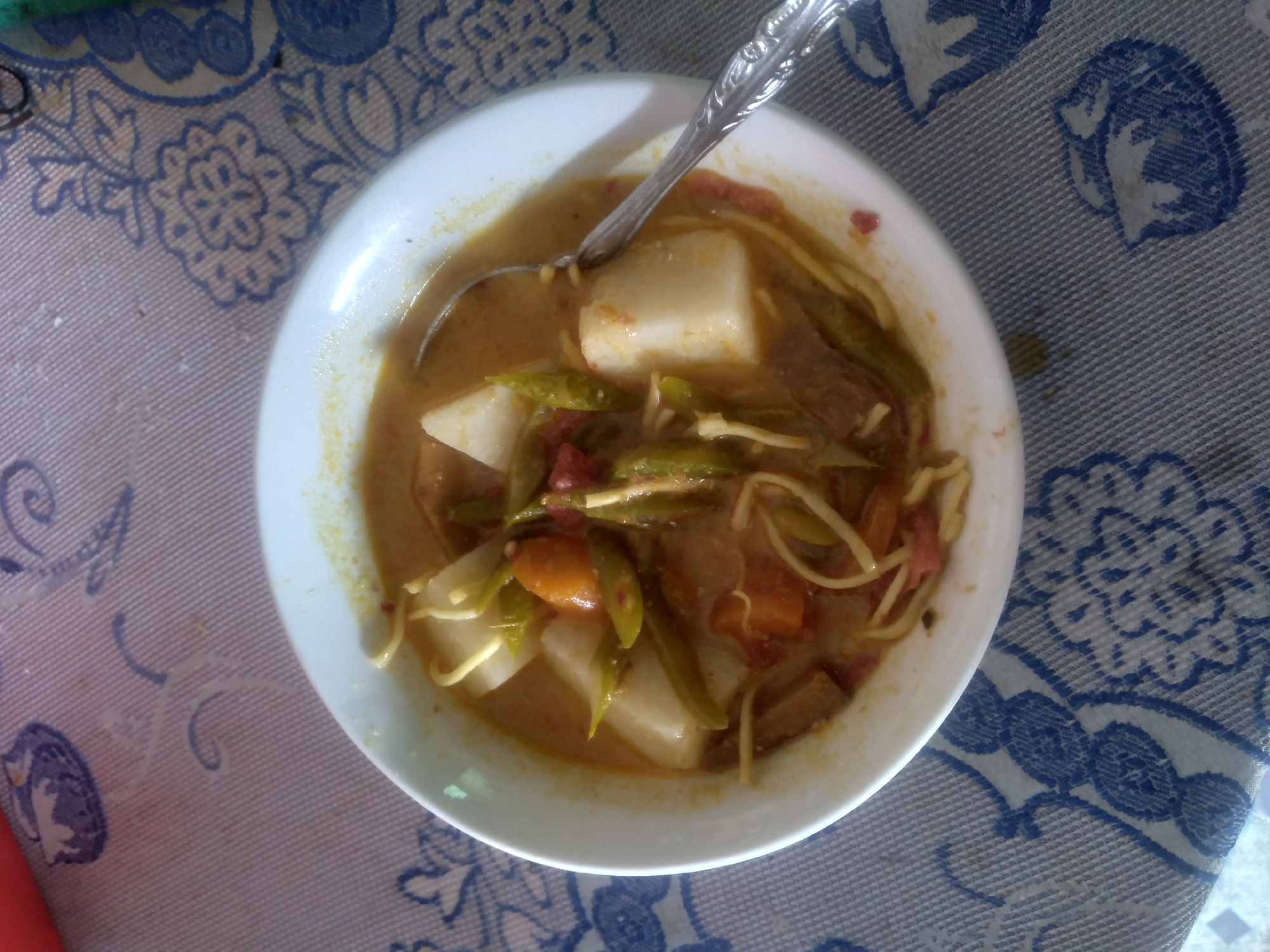 A white ceramic bowl of Lontong Sayur is placed on a blue and white patterned tablecloth. The bowl contains rice cakes (lontong), chayote, green beans, and shredded chicken in a savory coconut milk broth. A spoon rests in the bowl. The scene is likely from a home kitchen or a local eatery in Pekanbaru, Indonesia, given the dish's popularity in the region.