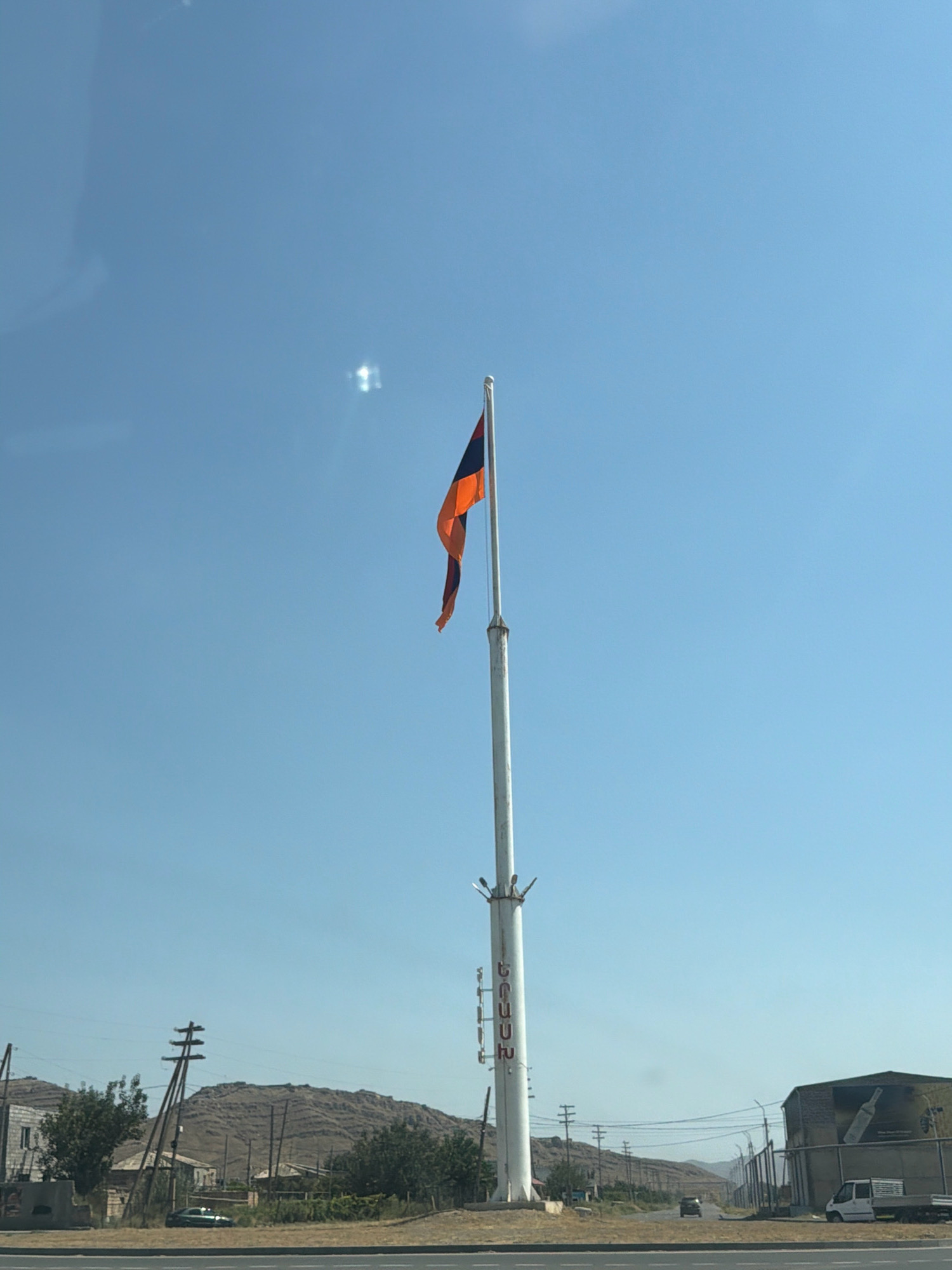 In the city of Yeraskh, Armenia, on a clear, sunny day with a bright blue sky, a large white flagpole stands tall in the foreground, flying the Armenian flag. The flag, with its horizontal bands of red, blue, and orange, billows gently in the breeze. The flagpole itself is adorned with what appear to be decorative elements and possibly lighting fixtures.
To the left of the flagpole, a distant, barren mountain range forms the backdrop. The landscape also includes scattered buildings, some appearing to be older, with visible architectural details, and others more industrial in appearance. Electrical poles and lines are present, indicating infrastructure in the area. In the middle ground, a road is visible, with a few vehicles, including a dark-colored car and a white van with a trailer, suggesting some level of activity. Trees and vegetation dot the landscape, with the overall environment appearing somewhat dry and arid. The image seems to have been taken from a moving vehicle, indicated by a slight glare or reflection on the left side of the frame, possibly from a car window.