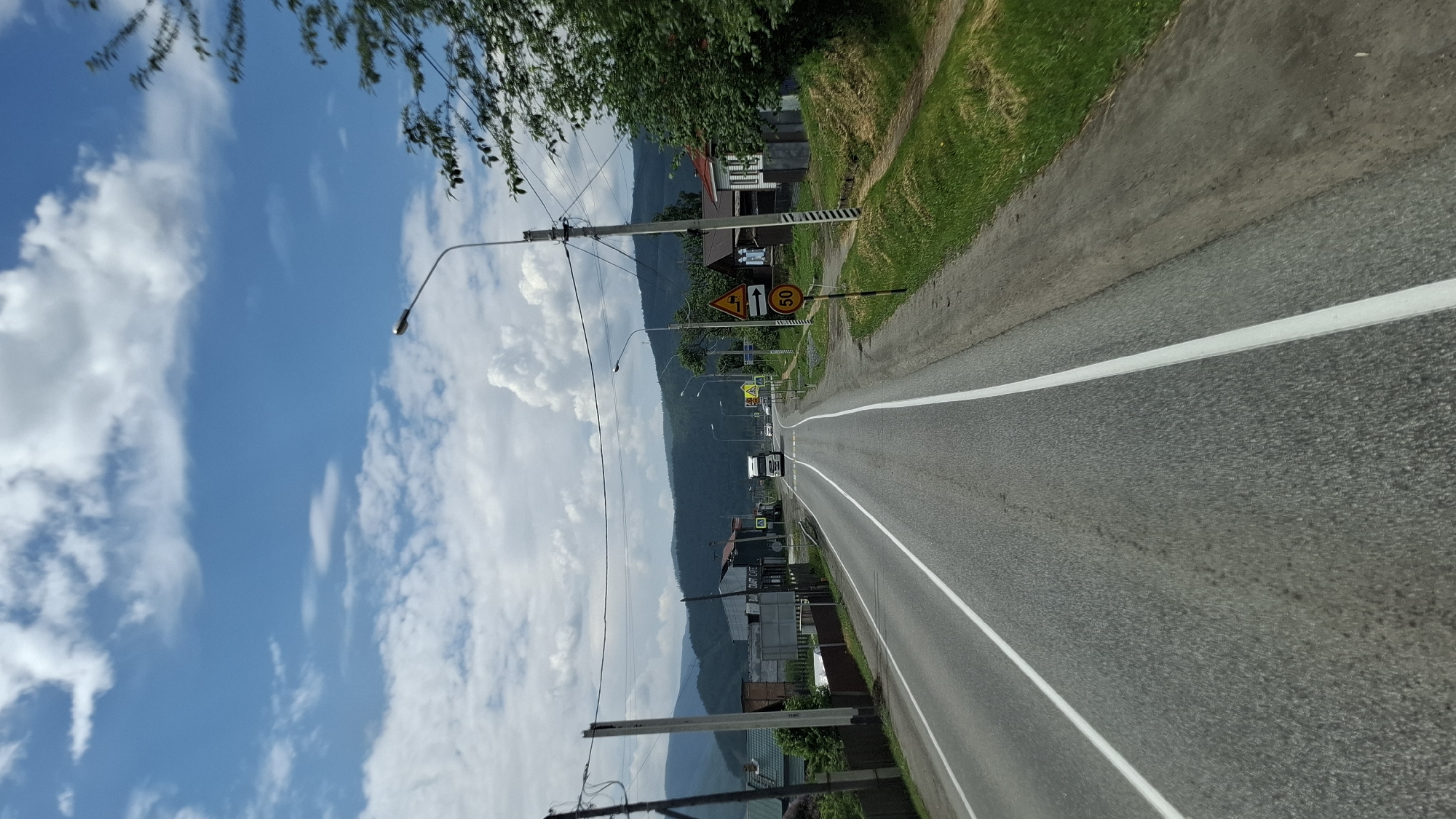 A wide, asphalt road with white lane markings leads through a rural area. On the right side of the road, a grassy shoulder gives way to a dirt path and then several houses. One house has a red roof and white siding. Further down the road, a speed limit sign indicates 50, and another sign shows a truck symbol and an arrow pointing right. On the left side of the road, there are more houses and buildings, including one with "CRAFT CAFE" visible on it. The road is lined with utility poles and wires. In the distance, rolling green hills and mountains are visible under a bright blue sky with scattered white clouds. A white truck is driving away from the camera in the distance. The scene appears to be in the village of Kultuk, Russia, likely during daylight hours.