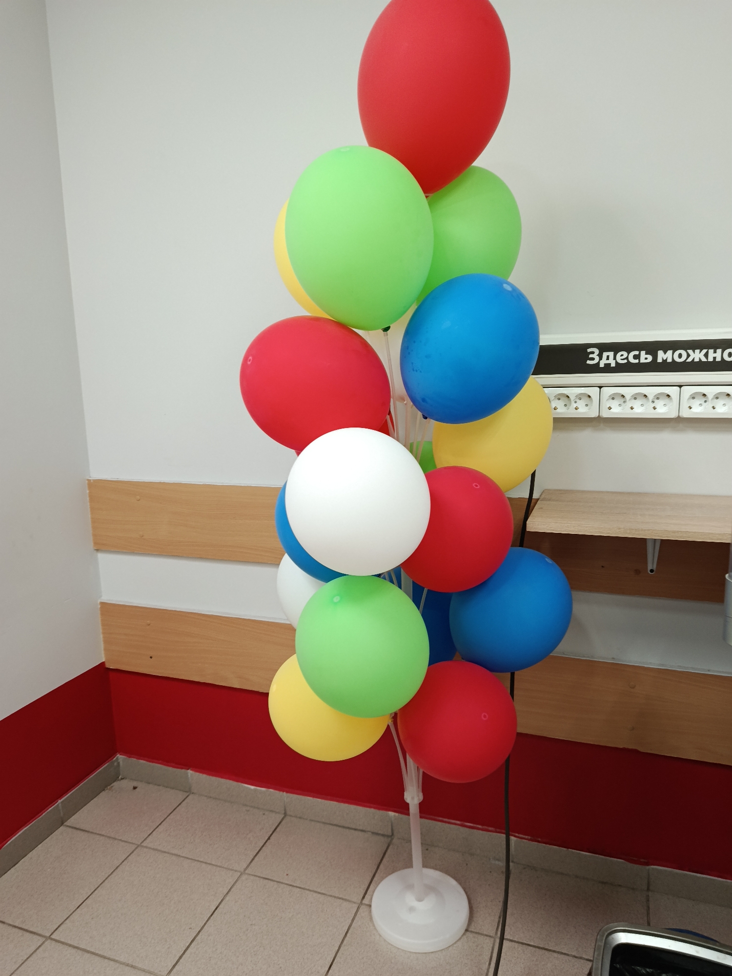 A cluster of colorful balloons is displayed on a stand. The balloons are arranged in a vertical column, with a red balloon at the top, followed by green, yellow, blue, red, white, blue, green, yellow, red, and blue balloons. The balloons vary in size and are all inflated. The stand is white and has a circular base.
The balloons are positioned in front of a light-colored wall with horizontal wooden panels. Above the wooden panels, there is a black strip with white text that reads "Здесь можно" which translates to "Here it is possible" or "You can here". Below the black strip, there are several white electrical outlets. A wooden shelf is mounted on the wall above the lower wooden panel.
The lower part of the image shows a tiled floor and a red baseboard. The overall scene appears to be indoors, possibly in a public space like an event venue or a store, given the electrical outlets and the suggestive text. There are no people visible in the image. The lighting is neutral, and there are no indications of the time of day or weather conditions. The arrangement of balloons suggests a festive or celebratory occasion.