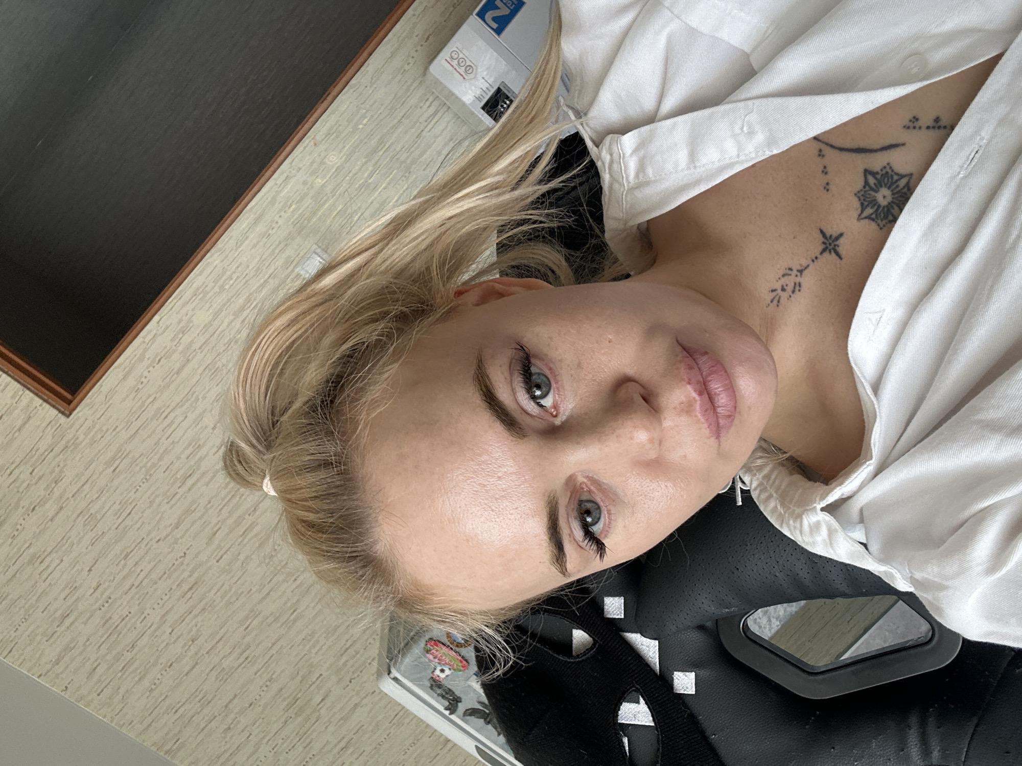 A woman with blonde hair in a ponytail is sitting in a chair. She is wearing a white shirt. She has tattoos on her chest. The background is a wall with a textured pattern.