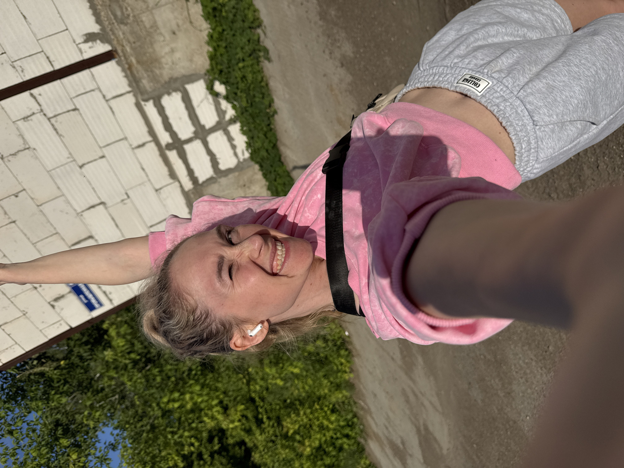 The image shows a young woman with blonde hair tied back, wearing a pink sweatshirt, gray shorts, and white wireless earbuds. She is outdoors in what appears to be a sunny day, judging by the shadows and bright lighting. The setting includes a paved pathway, a white brick wall, and lush green trees and foliage in the background. The woman is smiling broadly, with her eyes closed, and her arms are outstretched. She is holding the camera for a selfie, and her pose suggests a sense of joy or freedom. There is a black strap across her torso, possibly from a bag. A small blue sign with white text is visible on the brick wall. The label on her shorts reads "COLLEGE LEAGUE". The overall impression is a candid, cheerful moment captured outdoors.