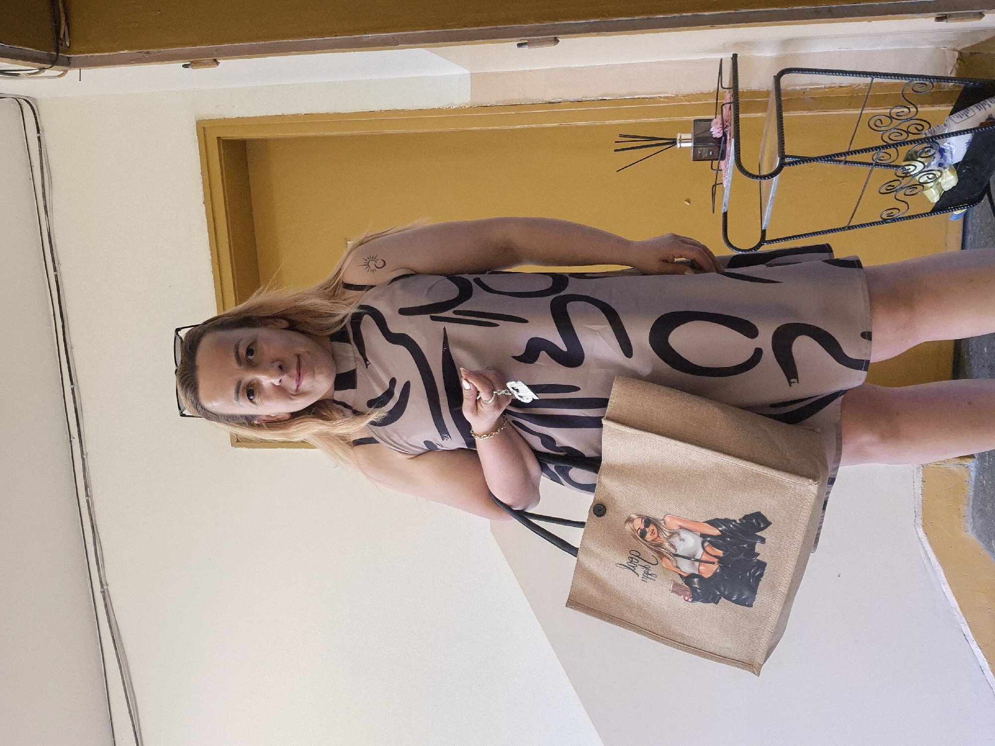 An adult female, with light skin and long blonde hair, stands in an indoor setting, likely a hallway or entrance. She wears a sleeveless, high-neck dress featuring a beige base with thick, abstract black curvilinear patterns. Sunglasses are perched on top of her head. Her face has light makeup, a small silver nose ring on her right nostril, and she looks directly forward with a slight smile. A small, stylized sun and crescent moon tattoo is visible on her left upper arm.
Her right hand holds a set of keys with a silver-colored, possibly heart-shaped, charm. Her left hand supports the black fabric handle of a large, light brown jute tote bag. The tote bag is printed with a graphic illustration of a blonde woman wearing sunglasses, a white crop top, and a black open jacket, holding a coffee cup, accompanied by the text "loop" and "yep!". She wears a gold-colored chain bracelet on her left wrist.
The background consists of plain off-white/cream walls. Along the top left wall, two dark electrical cables run horizontally, secured by white clips. To the right of the woman, a slender, black wrought-iron two-tiered stand is positioned. The top tier holds a dark diffuser bottle with reeds and a small pink artificial flower. The bottom tier, designed as a magazine rack, contains various plastic items. Behind the stand and to the right, a yellow-ochre painted door frame is visible, with a partially seen lighter beige door. Further to the right, another similar yellow-ochre door frame with a lighter beige door is partially visible. The floor is dark gray, and a yellow-ochre painted step or trim runs along the base of the left wall. The location is an indoor residential or commercial hallway in an unknown city.
