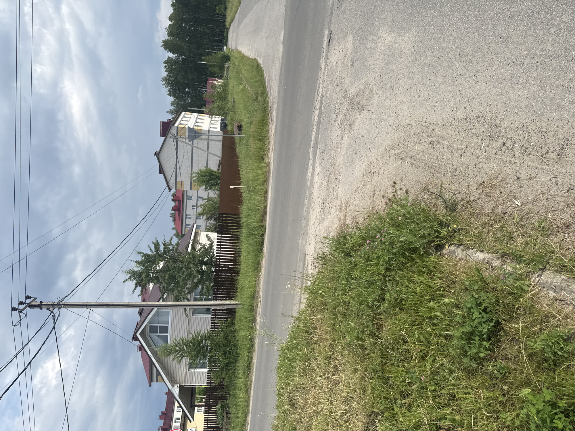 The image shows a street scene in Krasnye Baki, Russia, with residential buildings and a road. On the left, a light-colored house with a dark red roof and a triangular window is visible behind a wooden fence. Next to it is a multi-story building with a light-colored facade and a dark red roof. The street itself is paved, with a dirt and grass shoulder on the left. Overhead, power lines stretch across the frame, supported by a utility pole. In the background, a dense line of trees borders the right side of the road, and a white car is parked on the shoulder. The sky is overcast with gray clouds.