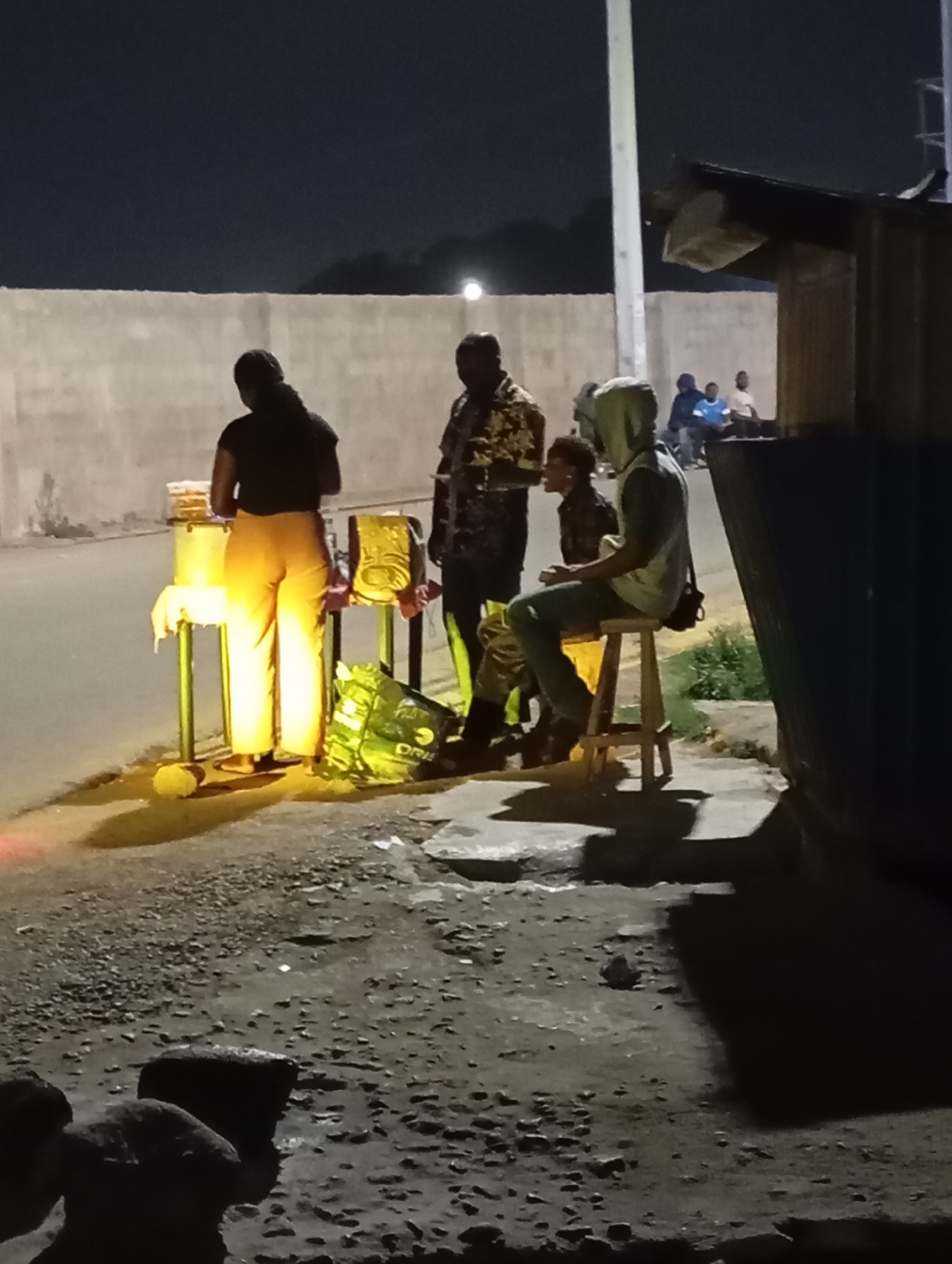 At night, near a wall and a small structure, several people gather at a street food vendor's stand in Jos, Nigeria. A person wearing yellow pants and a dark top is behind a table selling food; a man in a patterned shirt stands nearby. A seated person wears a hooded sweatshirt and jeans. Other individuals are indistinctly seen in the background, and bags are placed near the stand. The ground appears rough. The scene is illuminated by artificial light.