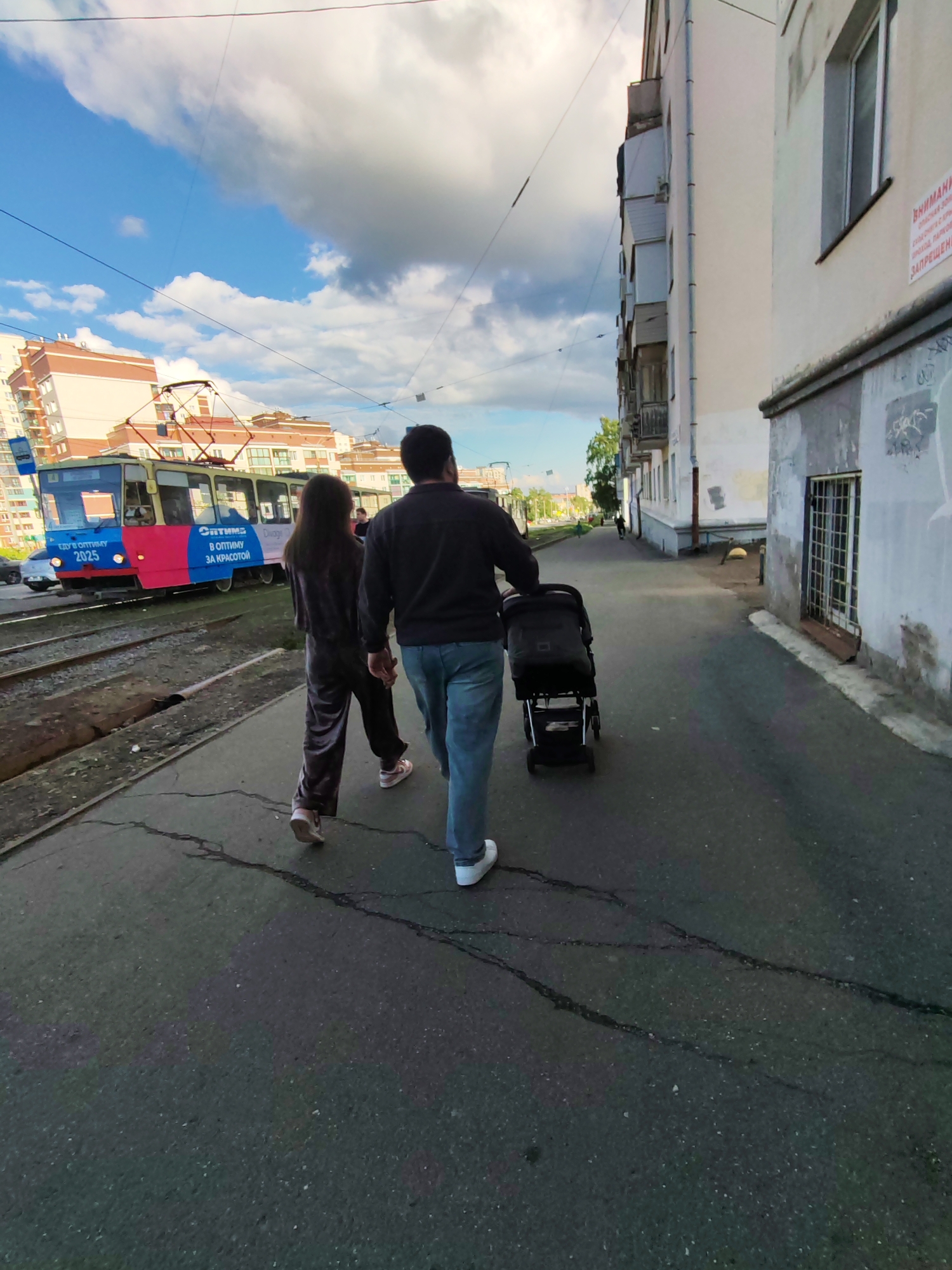 The image depicts an urban street scene in Izhevsk, Russia, on a bright, partially cloudy day, likely late afternoon or early evening. In the foreground, a couple is walking along a cracked asphalt sidewalk. The man, wearing a dark long-sleeved shirt, blue jeans, and white sneakers, pushes a black baby stroller. Beside him, a woman, dressed in a dark, possibly velvet, tracksuit and light-colored sneakers, walks.
To their left, parallel to the sidewalk, are tram tracks. A blue and red tram is visible on these tracks, with advertisements on its side. Visible text on the tram includes "ЕДУ В ОПТИМУ 2025" (I'm going to Optima 2025), "ОПТИМА В ОПТИМУ ЗА КРАСОТОЙ" (Optima to Optima for beauty), and "Chicago."
On the right, a multi-story building with a light-colored facade runs along the sidewalk. Its lower portion shows signs of wear, including peeling paint and graffiti. A barred window is seen at ground level. A red warning sign is affixed to the building, with the text: "ВНИМАНИЕ! ОПАСНАЯ ЗОНА. ОБРУШЕНИЕ С КРЫШИ. ПРОХОД, ПАРКОВКА ЗАПРЕЩЕНЫ" (Attention! Dangerous zone. Roof collapse. Passage, parking prohibited).
In the background, other residential buildings with varied architectural styles and colors are visible under a blue sky with scattered white clouds. Overhead wires, characteristic of tram lines, crisscross the frame. The scene captures a moment of daily life in an urban setting.