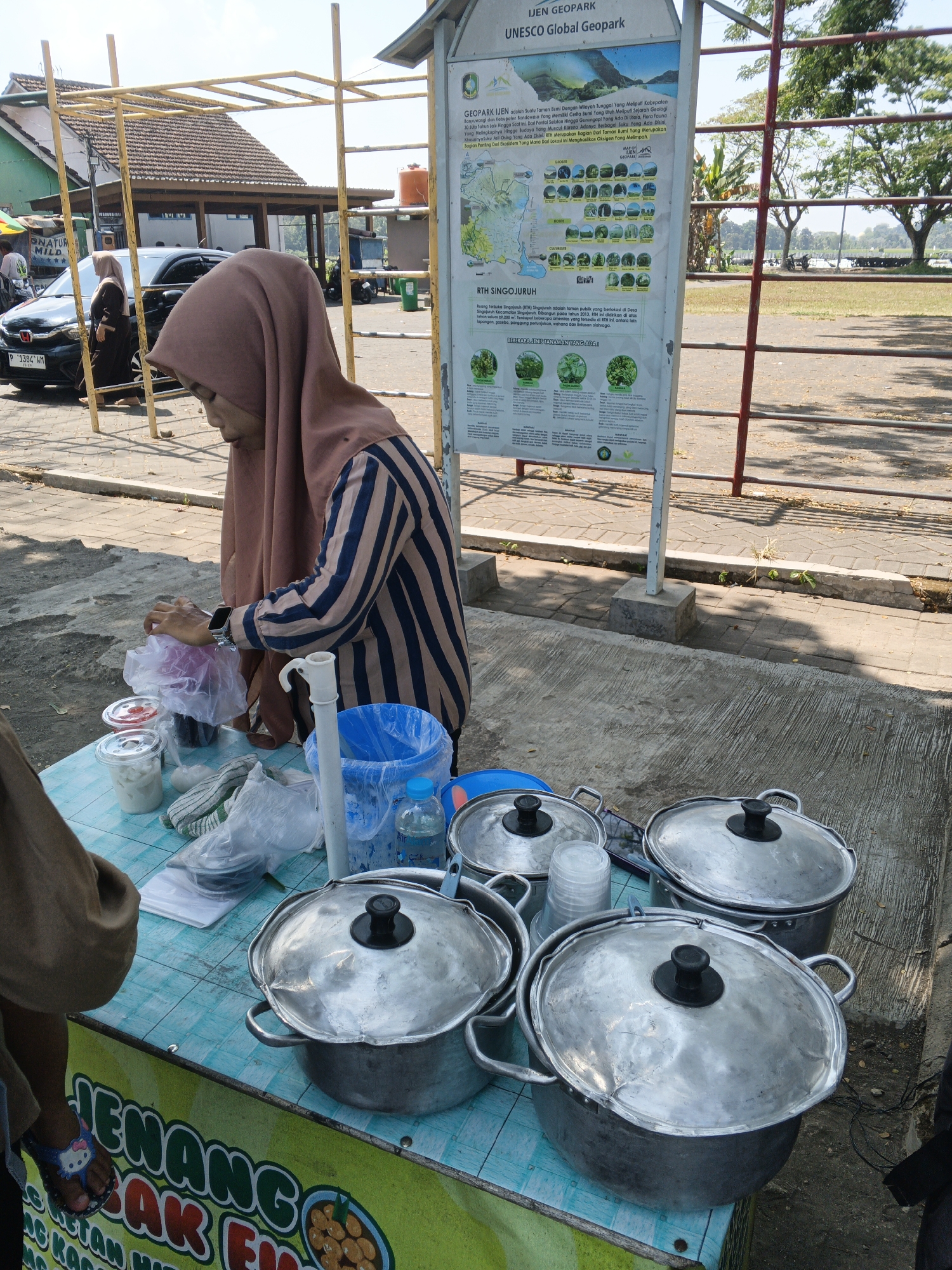 In Gumirih, Indonesia, a woman wearing a brown hijab and a striped shirt is seen preparing food at an outdoor stall. She is standing behind a turquoise table covered with various items, including stacked plastic cups, containers of food, and plastic bags. Several large, lidded metal pots are prominently displayed on the table, suggesting a food service operation. To the right of the table is a blue trash can and a bottle of water. In the background, a sign about the "IJEN GEOPARK UNESCO Global Geopark" stands tall, providing context for the location. Other elements in the scene include a yellow metal structure, a building with a tiled roof, a black car, and trees. The ground is paved with concrete.
