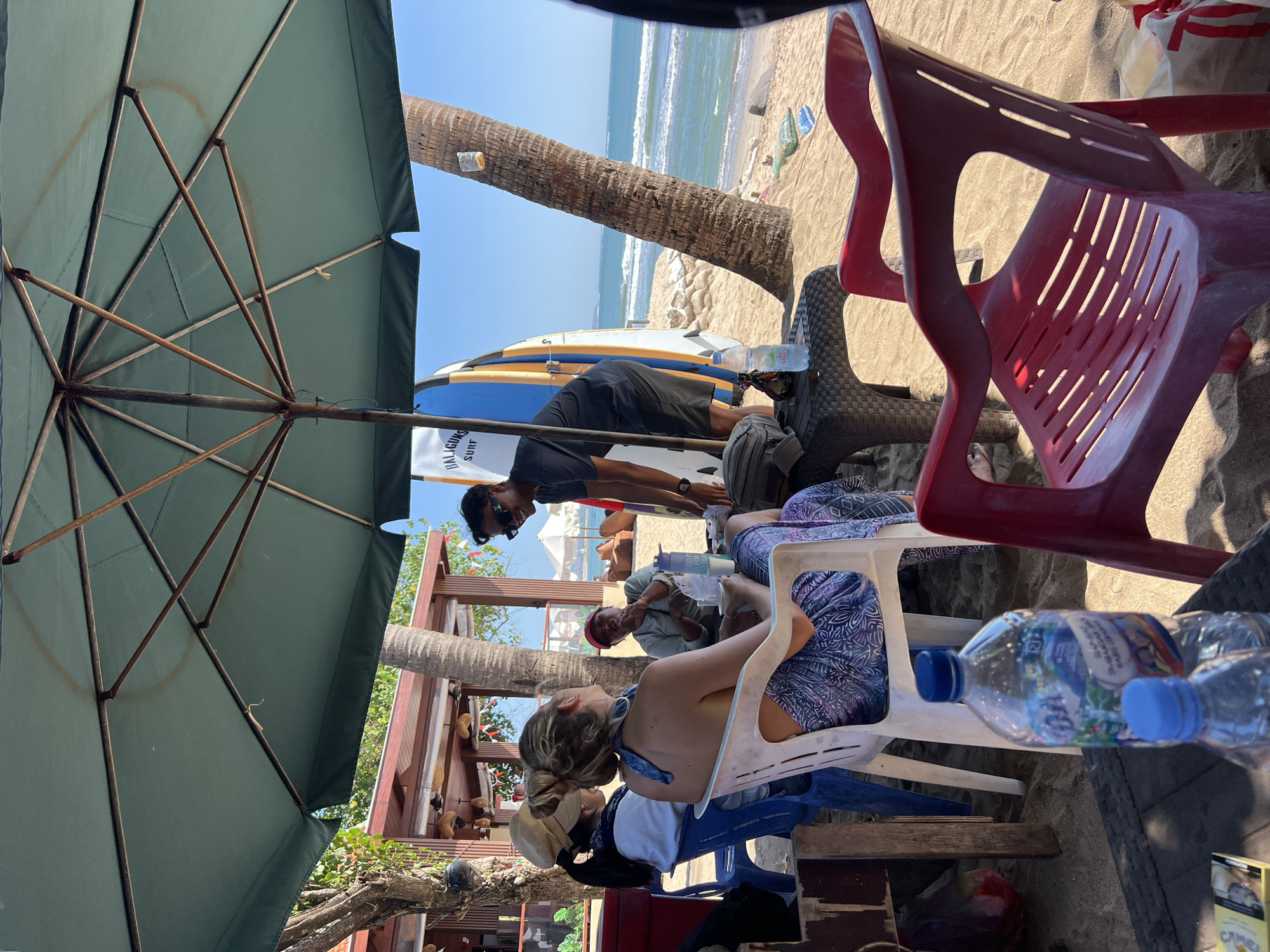 In Kuta, Indonesia, a group of people are seated under a large green umbrella on a sandy beach. A man in a dark t-shirt and shorts, wearing sunglasses, is interacting with a woman seated on a white plastic chair. The woman is wearing a blue and purple patterned dress and has her hair tied up. Another woman, wearing a light-colored cap and a white top, is seated to her left. A third woman in a red cap is seated behind them, appearing to be engaged in conversation. Two surfboards stand upright behind the group. Bottles of water and a pack of cigarettes are on a small table in front of the seated individuals. The ocean with gentle waves is visible in the background, with palm trees and a beachside structure to the left. The scene appears to be a relaxed gathering at a beachside establishment.