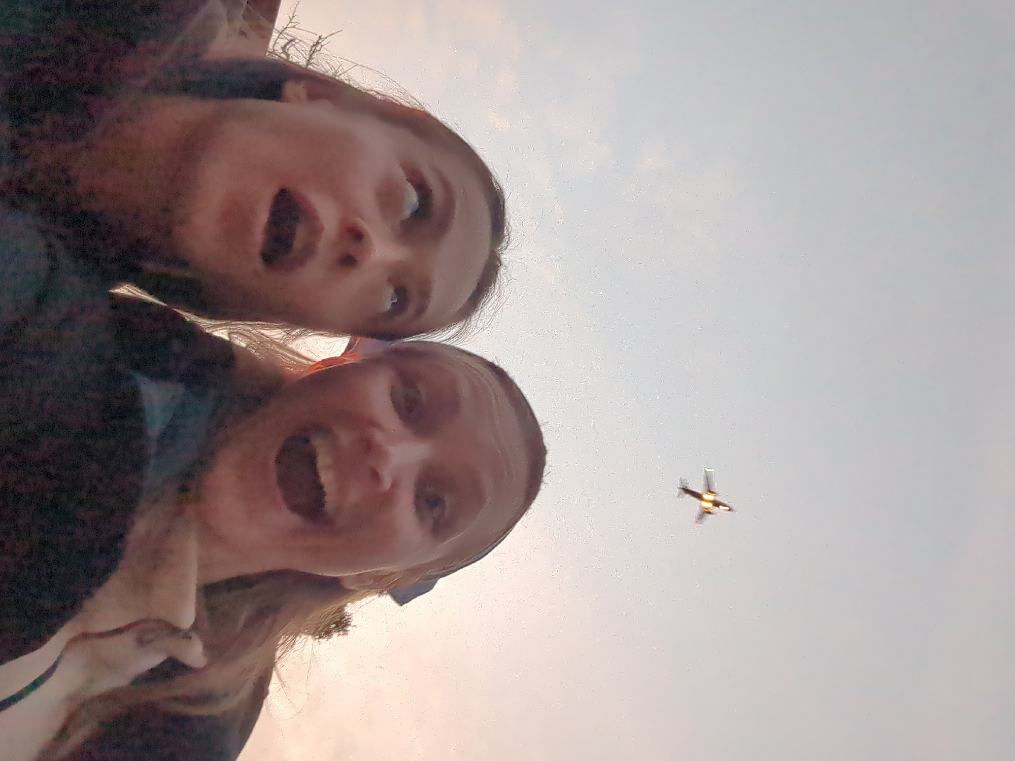 Two females are captured in a low-angle shot in Khimki, Russia, looking upwards towards the sky. The individual positioned on the left has dark hair, her head tilted back, eyes wide and directed upwards, with her mouth agape in an 'O' shape. The individual on the right has lighter hair, part of which is secured by a dark blue bow, her head also tilted back, eyes wide and directed upwards, with her mouth broadly open. Centered in the upper portion of the frame, a propeller-driven aircraft is visible, silhouetted against the sky but with prominent bright reflections on its fuselage and wings from direct light. The sky transitions from a light blue-grey at the top to a warm, pale orange-pink near the horizon, consistent with either dawn or dusk. Minimal, dark foliage is faintly discernible on the far right edge of the frame.