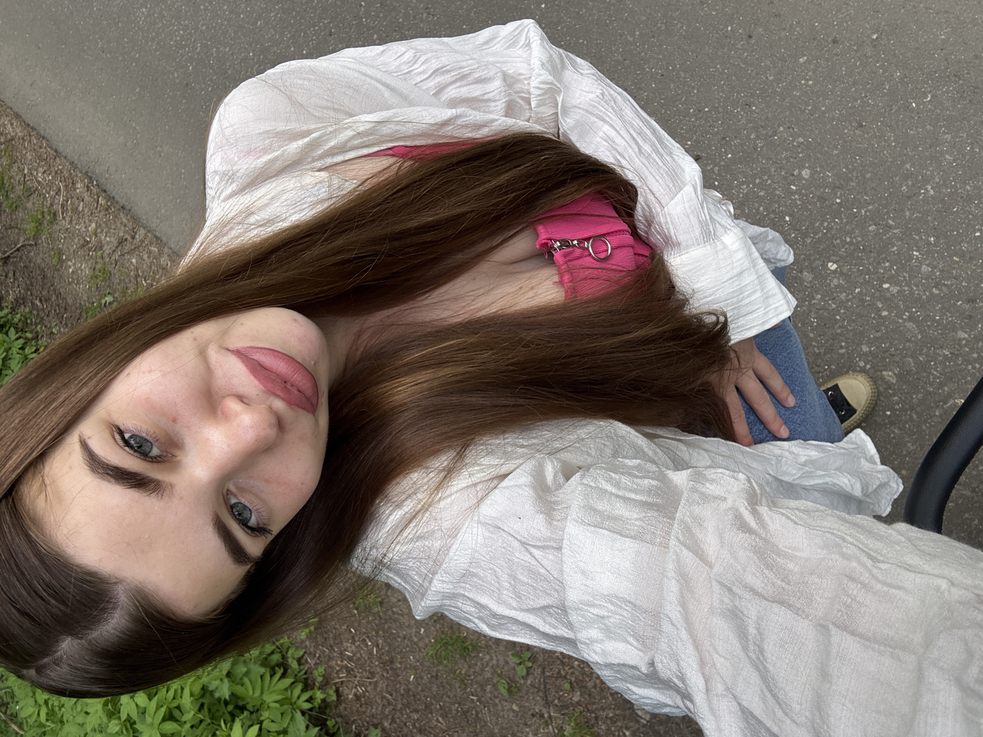 A young woman with long brown hair and blue eyes is the primary subject of this tilted selfie. She is wearing a white crinkled shirt over a pink top with silver hardware. The image also captures her blue jeans and a partially visible tan and black shoe.
The background is a mix of urban and natural elements. To the right of the woman is paved asphalt, likely a street or sidewalk. To the left is some green vegetation, possibly grass or small plants. A black curved object is also visible on the right, possibly a handle or part of a structure. There's no evidence in the picture that it was taken in Мещёры, Russia.