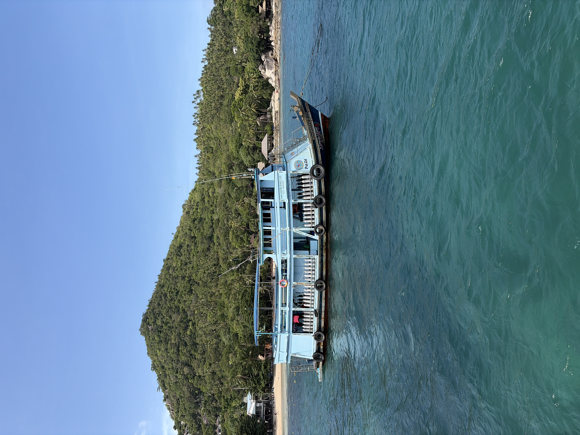 In the foreground, the turquoise waters of the Gulf of Thailand gently ripple under a clear blue sky. A light blue dive boat, equipped with numerous scuba tanks lined up along its side, is anchored offshore. The boat displays the text "PADI" and a website address. In the mid-ground, a sandy beach with scattered beach chairs and a few buildings is visible, backed by a densely forested, green hillside. Palm trees are interspersed throughout the vegetation, and several large boulders are present near the shoreline. The scene is characteristic of the coastal areas in Ko Pha-ngan District, Thailand, likely a popular spot for diving excursions.