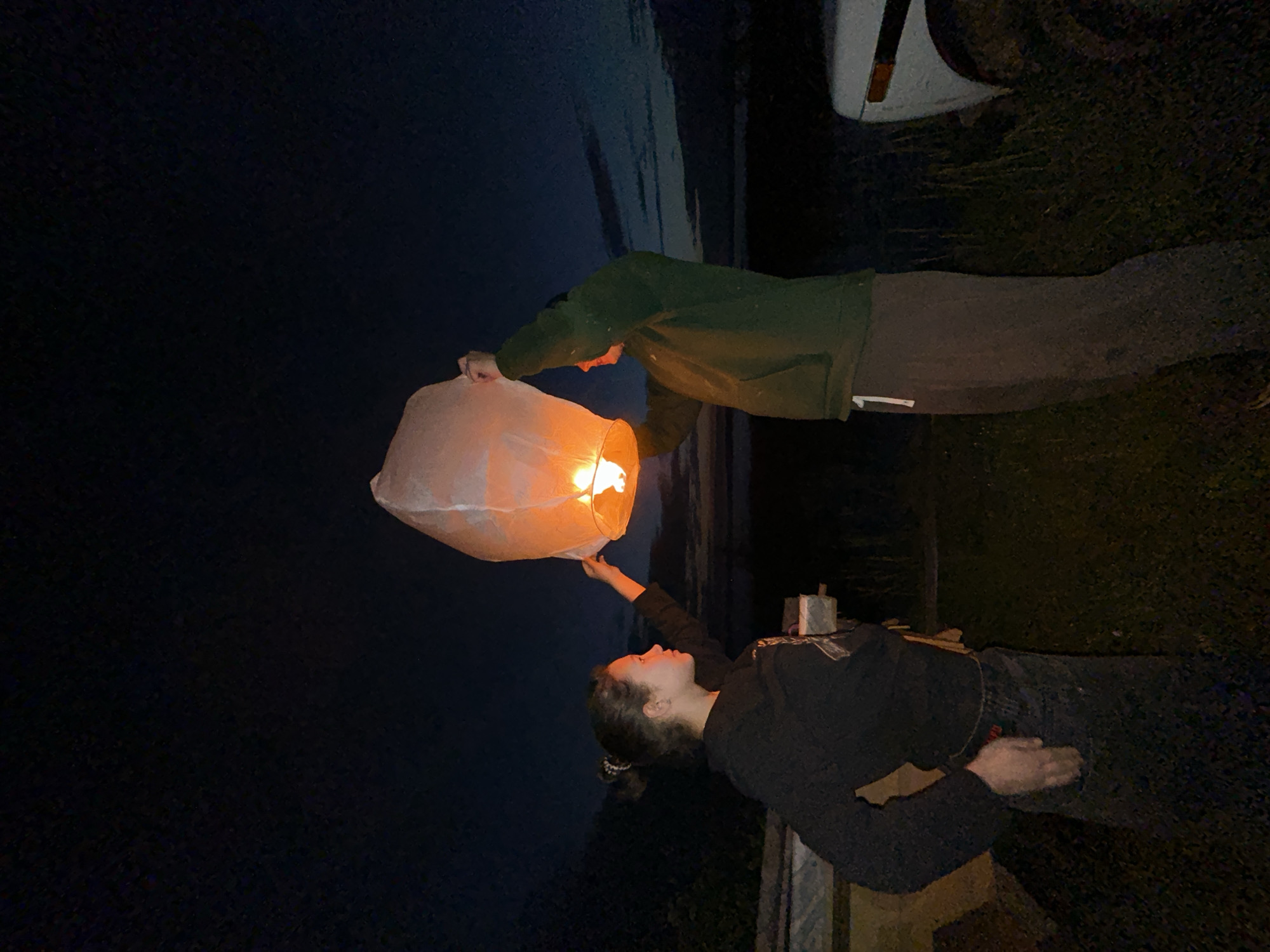 Two individuals, one female and one male, are outdoors at night, holding a lit sky lantern. The female, on the left, is wearing a black hooded sweatshirt and dark pants. The male, on the right, is wearing a green hooded sweatshirt and dark pants. They are standing on a grassy area, with a dark, cloudy sky overhead. To the left of the individuals, there is a structure that appears to be a wooden fence or shed. To the right, a portion of a white vehicle is visible. The scene is located in Plishkino, Russia.