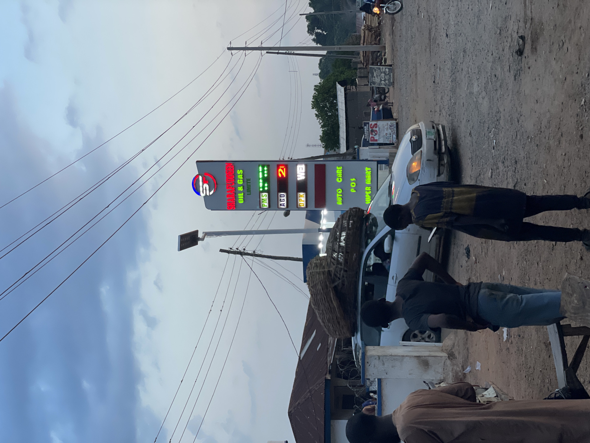 The image depicts a street scene in Akwanga, Nigeria, featuring a Sharafuddeen Oil & Gas Limited gas station. The gas station has a large sign displaying fuel prices (PMS, AGO, OPK), alongside other services like Auto Care, POS (Point of Sale), and a Super Mart. The prices on the LED sign read "21". Several people are in the foreground, appearing to be interacting or standing near a parked white car. Overhead, power lines stretch across the sky. In the background, there are other buildings, a motorbike, and what appears to be lumber. The setting suggests a commercial or roadside area.