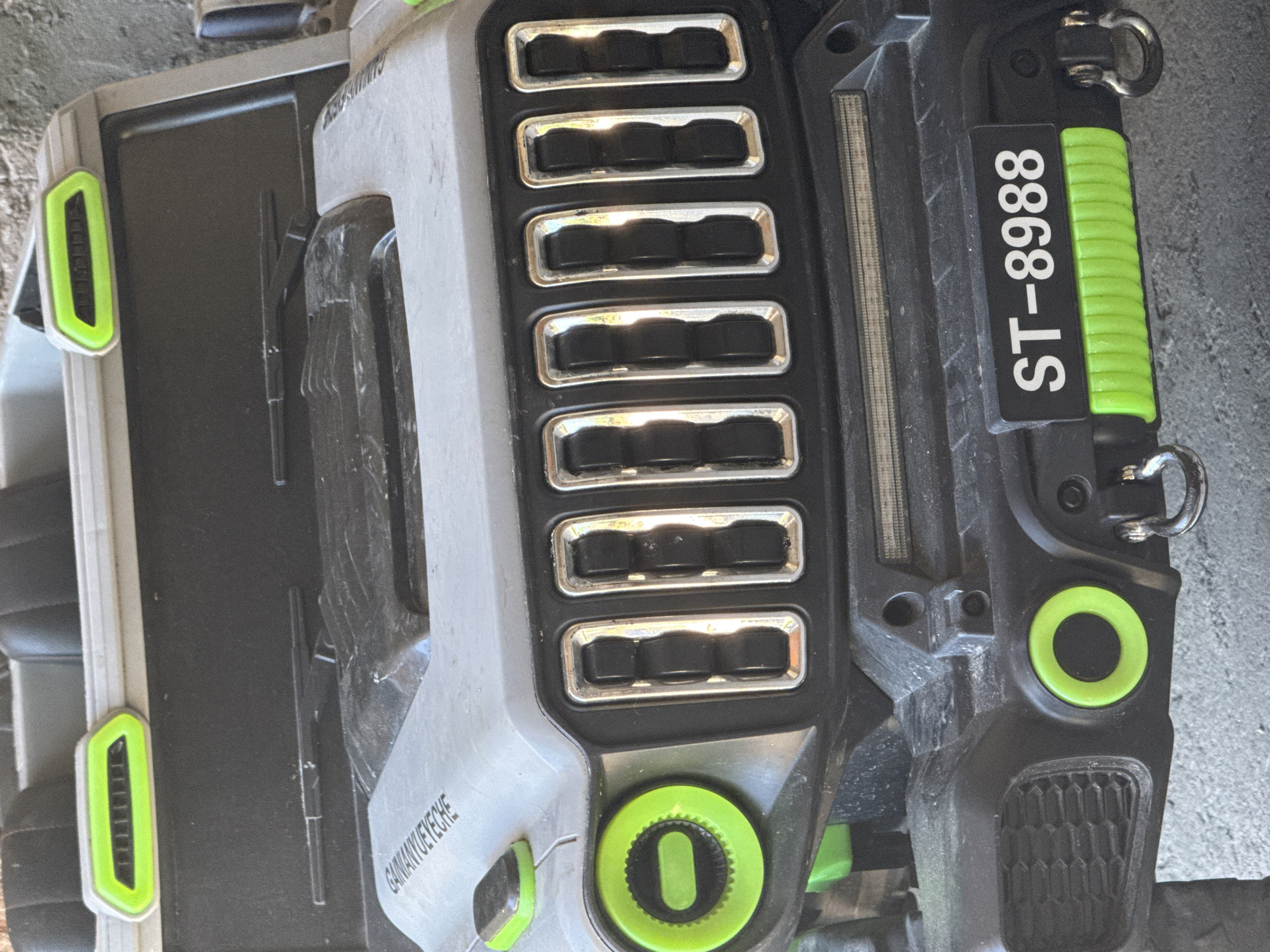The image shows a close-up of a piece of equipment, possibly a lawnmower or similar outdoor tool. It's predominantly gray and black with green accents. Notable details include a series of rectangular vents with metallic borders, a label that reads "ST-8988", and several circular green elements. The equipment appears to be resting on a concrete surface. The location is not discernible from the image.