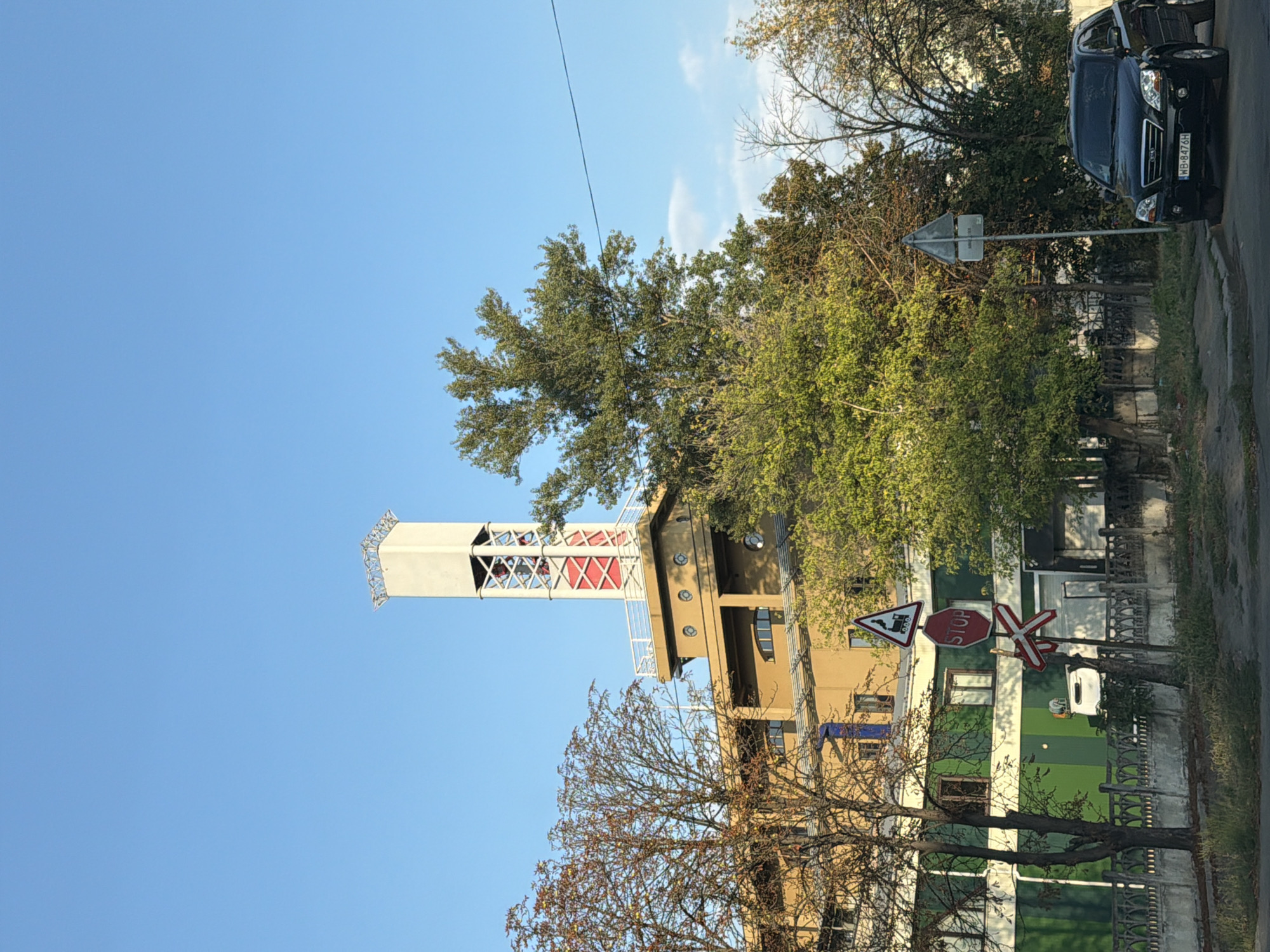 In the city of Kyiv, Ukraine, a multi-story building with a distinctive tower is visible against a clear blue sky. The building is painted in shades of yellow and green. A tall, square tower with a white top section stands out, featuring red lattice-work visible through openings. Trees with varying degrees of foliage, some displaying autumn colors, surround the structure. In the foreground, a black SUV is parked on the side of a street. Traffic signs, including a stop sign and a railway crossing sign, are present near the road. A concrete fence runs along the edge of the property.