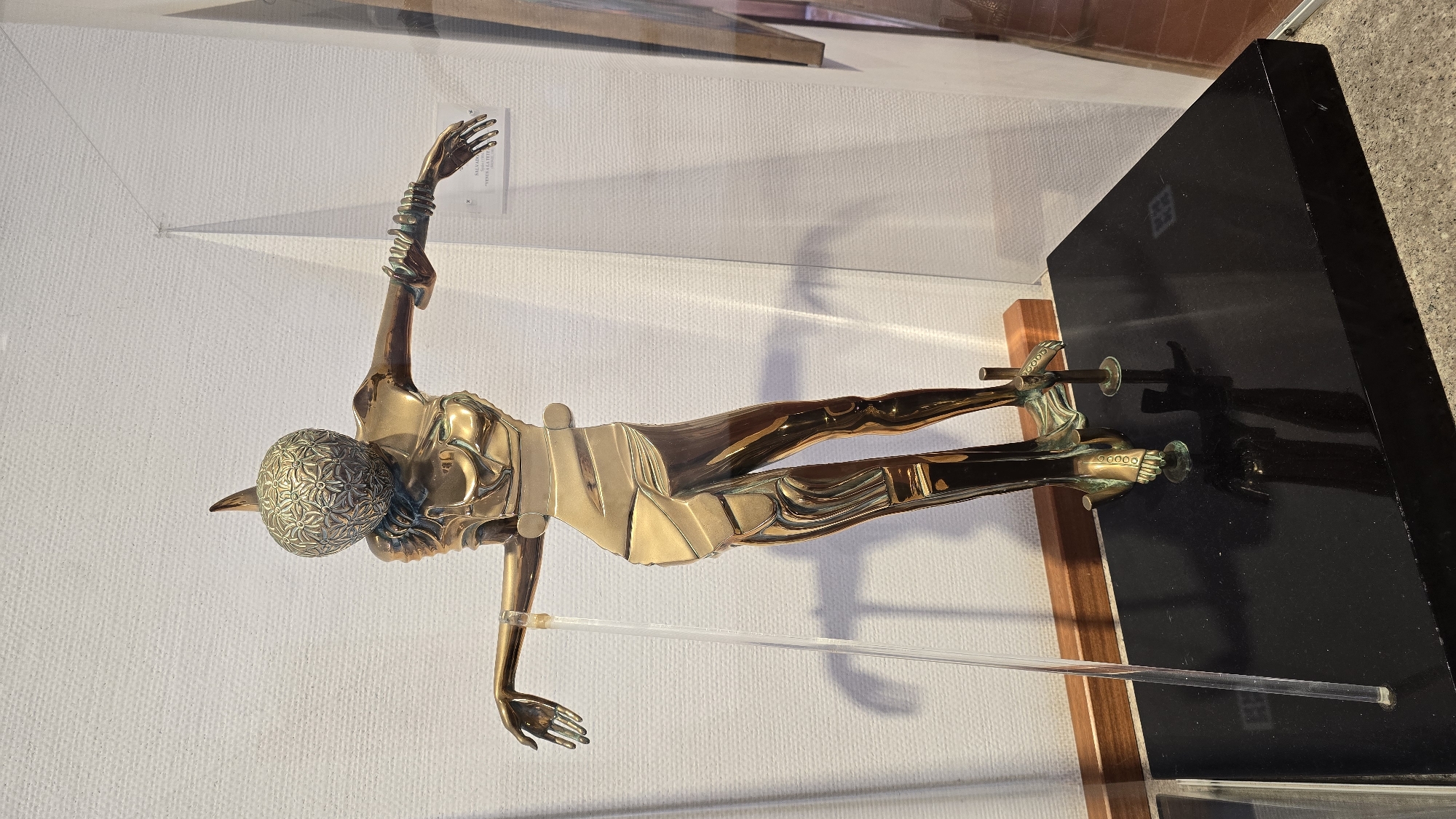 A bronze sculpture of a disembodied torso with legs is displayed on a black pedestal. The sculpture is a metallic gold color and has a detailed, fragmented design. The head is spherical with a patterned surface and a pointed protrusion. The torso is slender and appears to be made of interlocking metallic pieces. The arms are extended outwards, and the hands are open. The legs are thin and jointed, with feet that are also detailed. The sculpture is positioned in front of a white, textured wall and is partially enclosed by a glass display case. A clear acrylic rod is positioned to the left of the sculpture, and a small plaque with text is visible on the wall behind the sculpture. The scene is observed from a slightly elevated perspective, looking down at the sculpture. Based on available information, this sculpture is located in the Dali Museum in the city of Hof HaCarmel Regional Council, Israel.