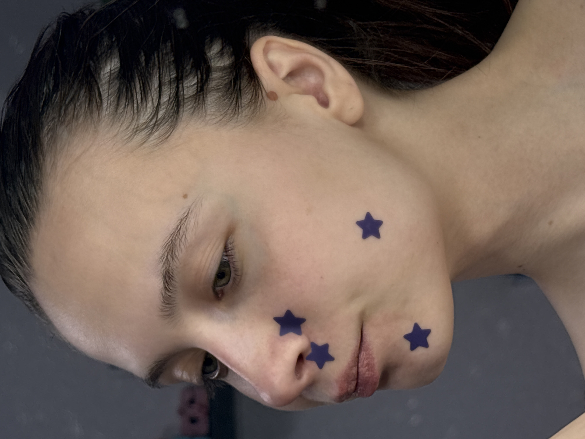 The image is a close-up portrait of a young woman with fair skin and dark hair that appears wet or styled with product, giving it a slicked-back look. Her head is tilted downward and slightly to the left. She has several dark blue star stickers on her face. There is a mole visible on her right ear. The background is blurred and appears to be a wall, but it is not possible to confirm this. There is no information in the image itself to indicate a location in the city of Прикамский, Russia.
