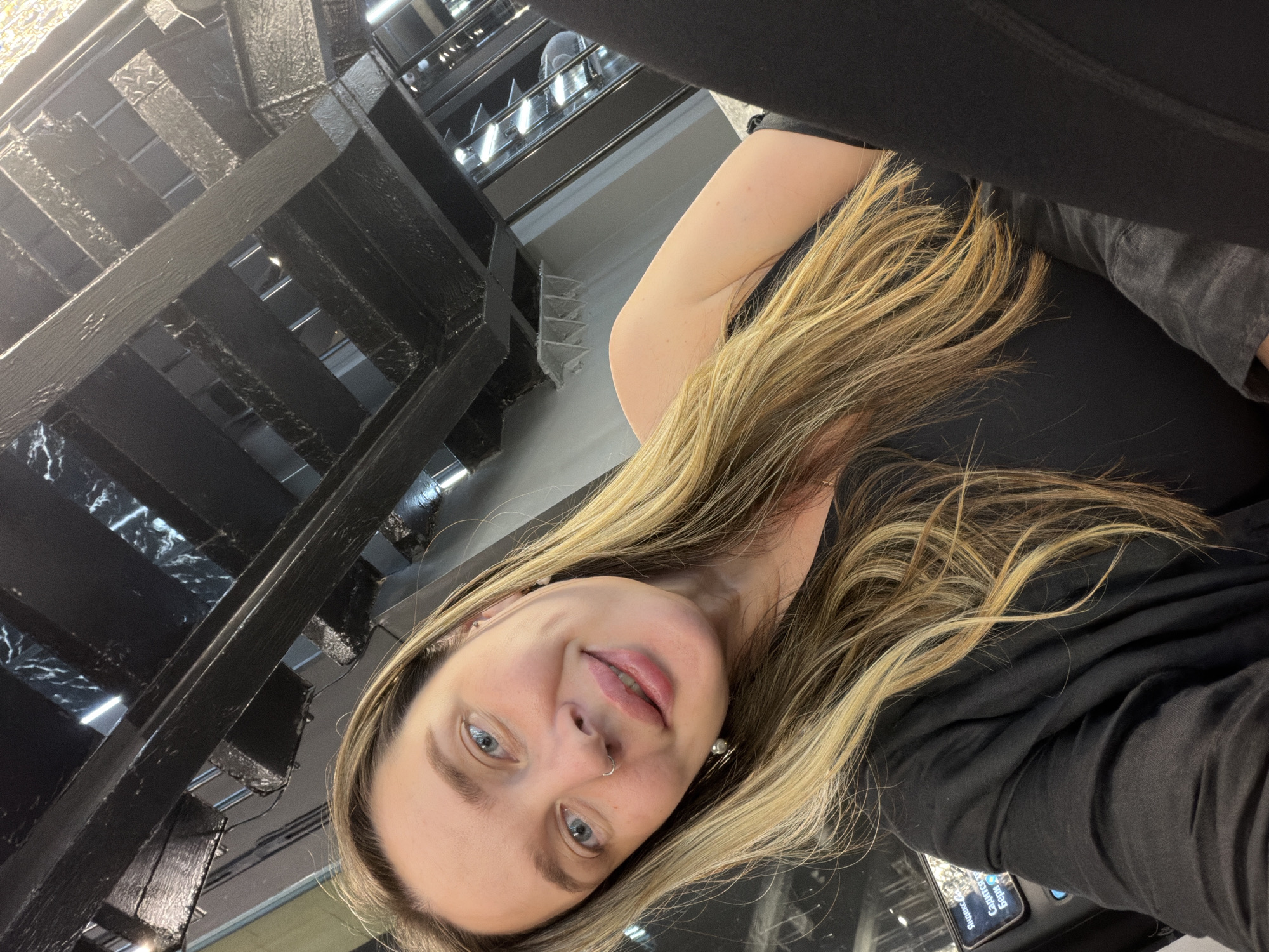 A woman with blonde hair and blue eyes is posing for a photo. She is wearing a black shirt and has a nose ring. Behind her is a black staircase. The location is Moscow, Russia.