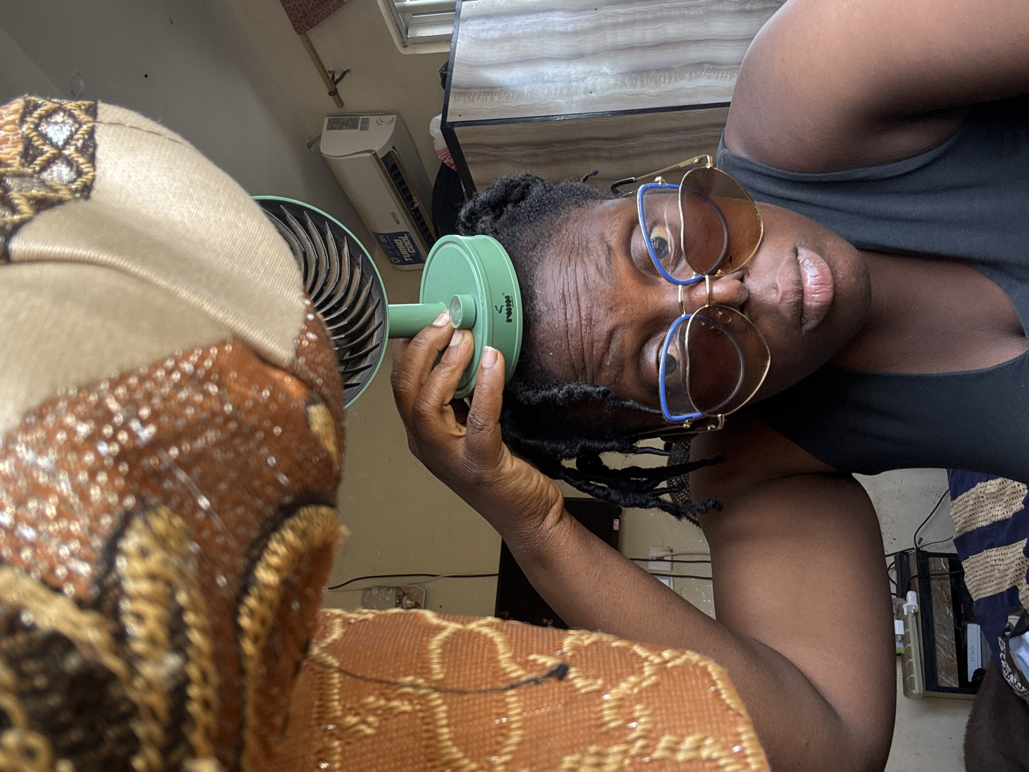 A medium shot captures a Black woman positioned indoors, indoors in Jawu, Nigeria. She is looking directly at the camera with a neutral expression, wearing large, brown, heart-shaped sunglasses with blue frames, and a black tank top. Her dark hair is styled in dreadlocks. The woman is balancing a small, green, electric fan on top of her head with her right hand. She is seated, and the camera is positioned slightly below her, looking upwards. In the foreground, an out-of-focus, richly textured, brown and gold fabric drapes from the top left of the frame. The background reveals an indoor environment with a visible air conditioning unit mounted on the wall, a wooden wardrobe, and a bedside table with electronic devices. The lighting suggests it is daytime.