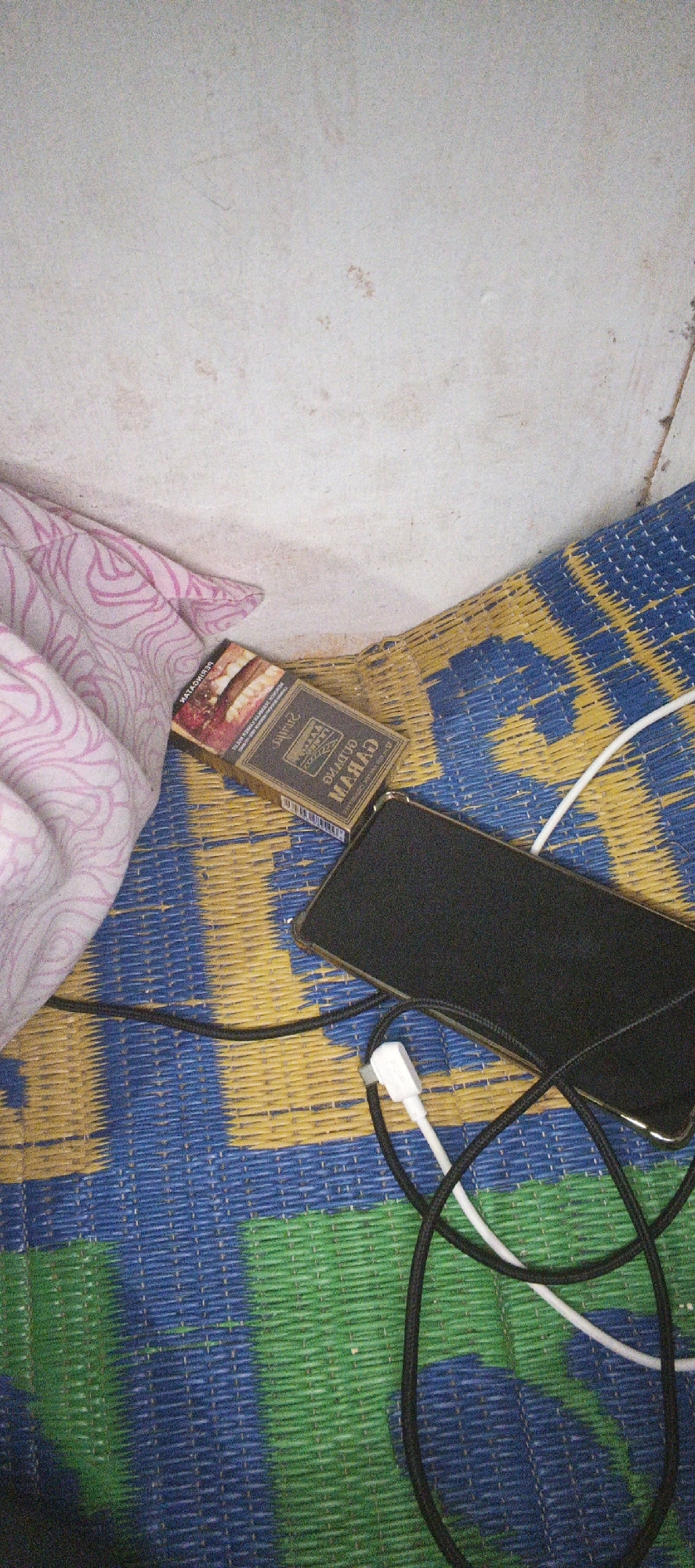 A flat lay image shows a cigarette pack and a smartphone on a woven mat. The cigarette pack, oriented diagonally, is gold and red with text in Indonesian. A black smartphone, lying flat, is positioned to the right of the cigarette pack. Two charging cables, one white and one black, are connected to the smartphone and extend across the mat. The mat is blue and yellow, with some green sections. A pink and white patterned fabric is draped on the left side of the image, partially obscuring the cigarette pack. The background is a textured, off-white wall. The scene appears to be indoors, with no discernible elements to pinpoint a specific location within Cinta Dame, Indonesia.