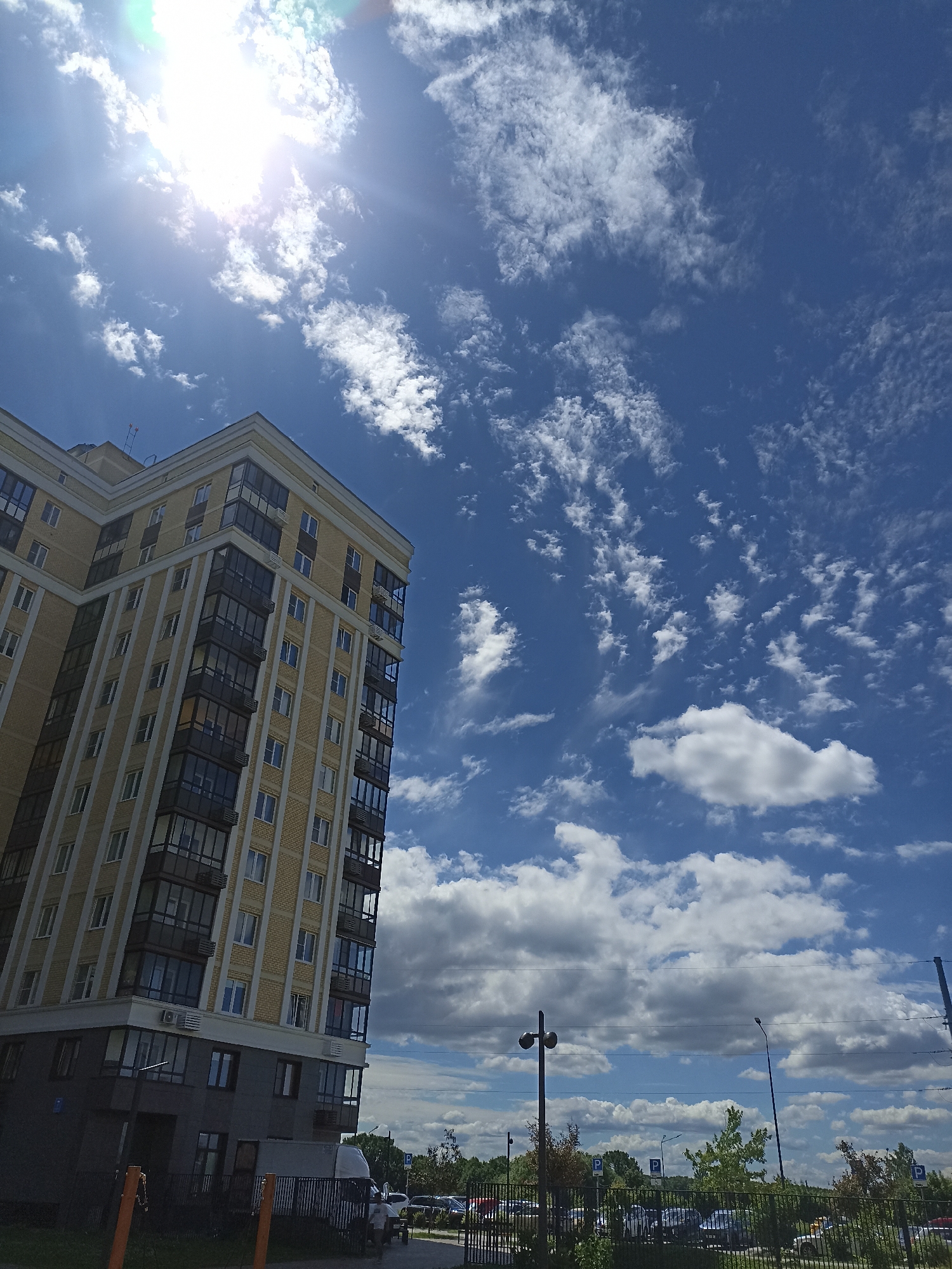 An exterior, medium-angle, low-light, low-saturation, full-day shot captures a modern, beige-brick apartment building in Ostafievo, Russia, under a bright, partly cloudy blue sky. The sun is visible in the upper left corner, casting a strong glare. The building features numerous windows and balconies. In the foreground, a black metal fence separates the building from a parking lot filled with cars. A white van is parked in the lot. The scene appears to be a typical urban environment.