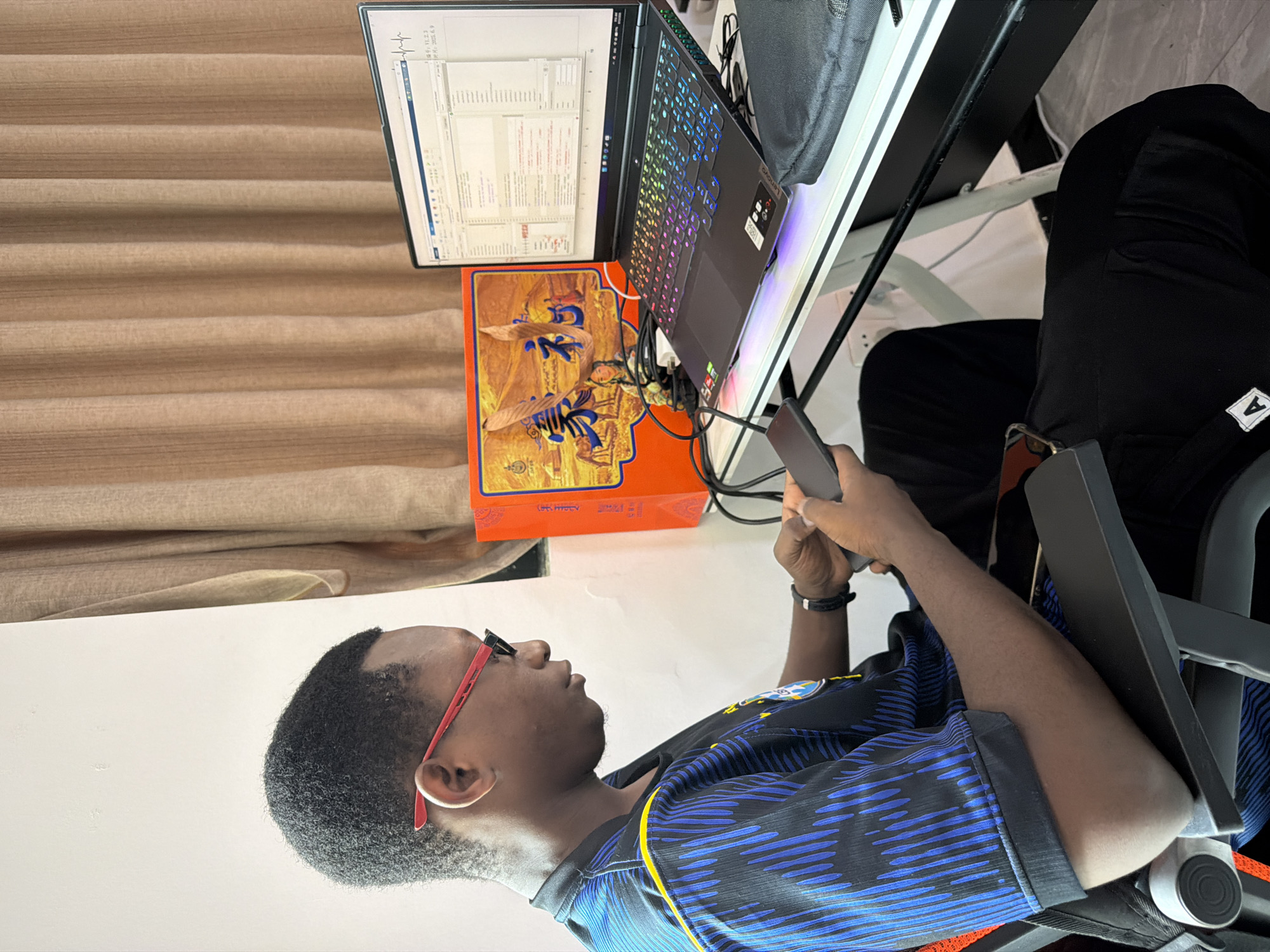 A young Black male wearing red-rimmed glasses and a blue and black striped shirt is seated in a black gaming chair, looking down at a smartphone he holds in his hands. He is positioned to the left of a desk. On the desk, to his right, is an open laptop with a colorful, illuminated keyboard, displaying a software interface. Beside the laptop is a bright orange box with an image of a pastoral scene featuring animals and people, and some text. A beige curtain hangs in the background, covering a window. The scene appears to be an indoor setting, possibly a home office or gaming room. There is no information available to ascertain its location within the city of Jiangjin, China.