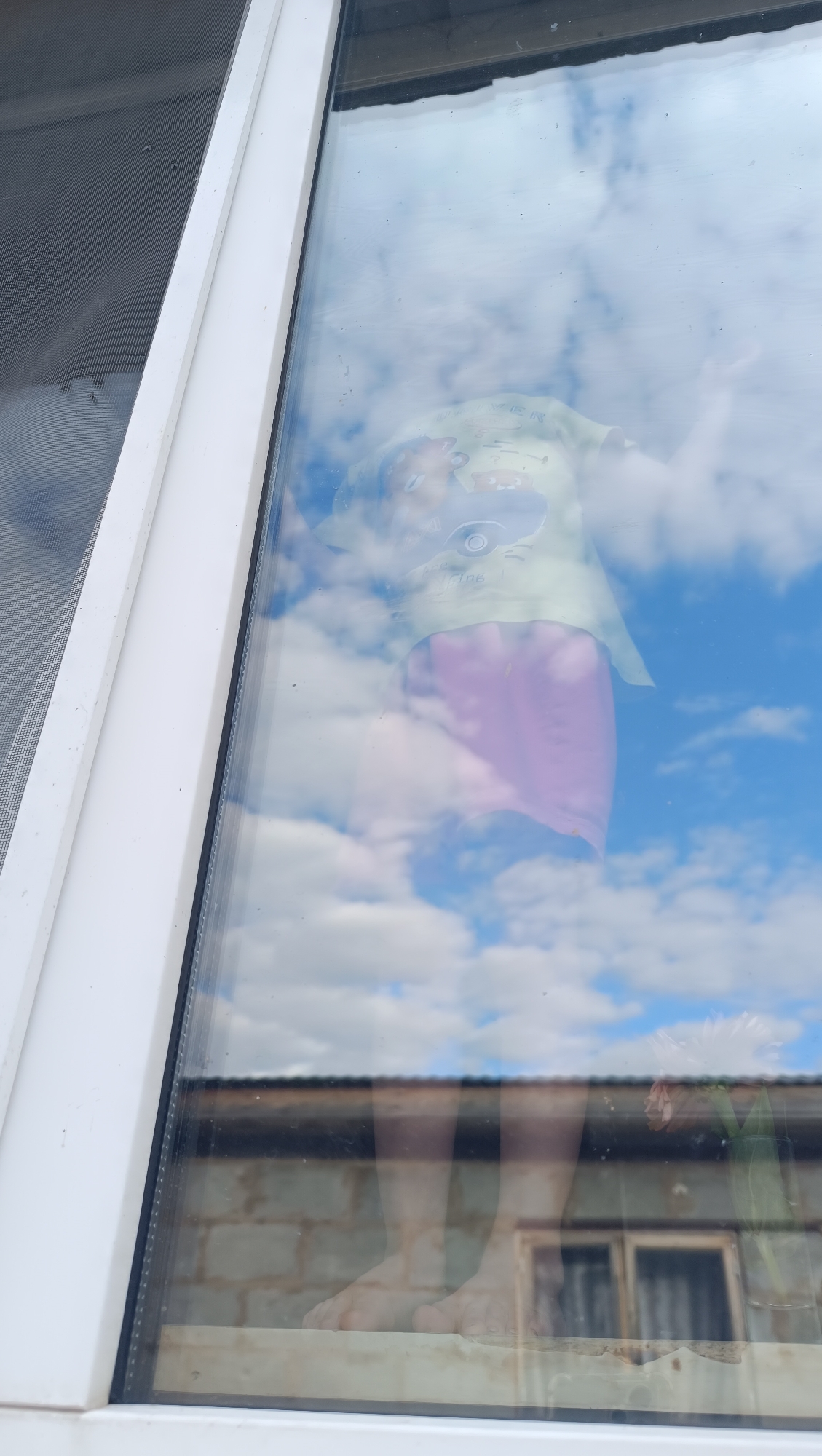 The image, taken in Karsha, Russia, displays a reflection on a windowpane. The reflection shows a child wearing a light green shirt with graphics and purple shorts, standing with their arms outstretched. The background of the reflection consists of a blue sky with scattered white clouds. Through the window, the actual scene shows bare feet standing on a wooden surface. Behind that, a brick wall and a framed object are visible. A vase with a flower is also reflected on the right side of the window. A screen is visible on the left side of the window frame.