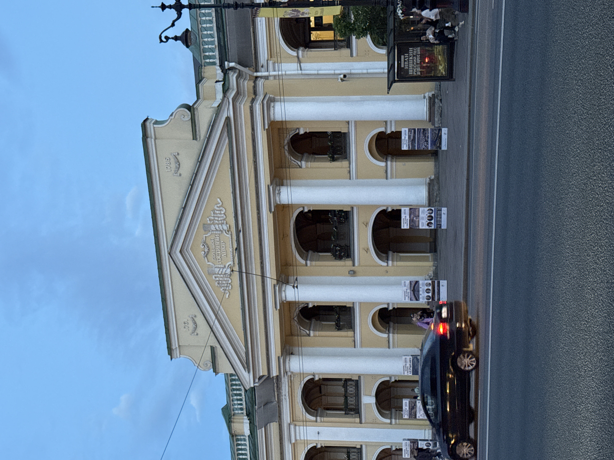 The image shows a street view in front of the historic Bolshoi Gostiny Dvor building in Saint Petersburg, Russia. The building, painted a pale yellow, features grand classical architecture with a prominent portico supported by large white columns, arched windows, and decorative friezes. The dates "1781" and "1785" are visible on the facade.
In the foreground, a black sedan is driving on a multi-lane asphalt road. The sky above is a dusky blue with some wispy clouds, suggesting it is late evening or early morning.
On the sidewalk in front of the building, several display stands with posters are set up. To the right, a bus stop shelter is visible with people sitting inside and standing nearby. The overall atmosphere is that of a city street scene during a time of low light.
