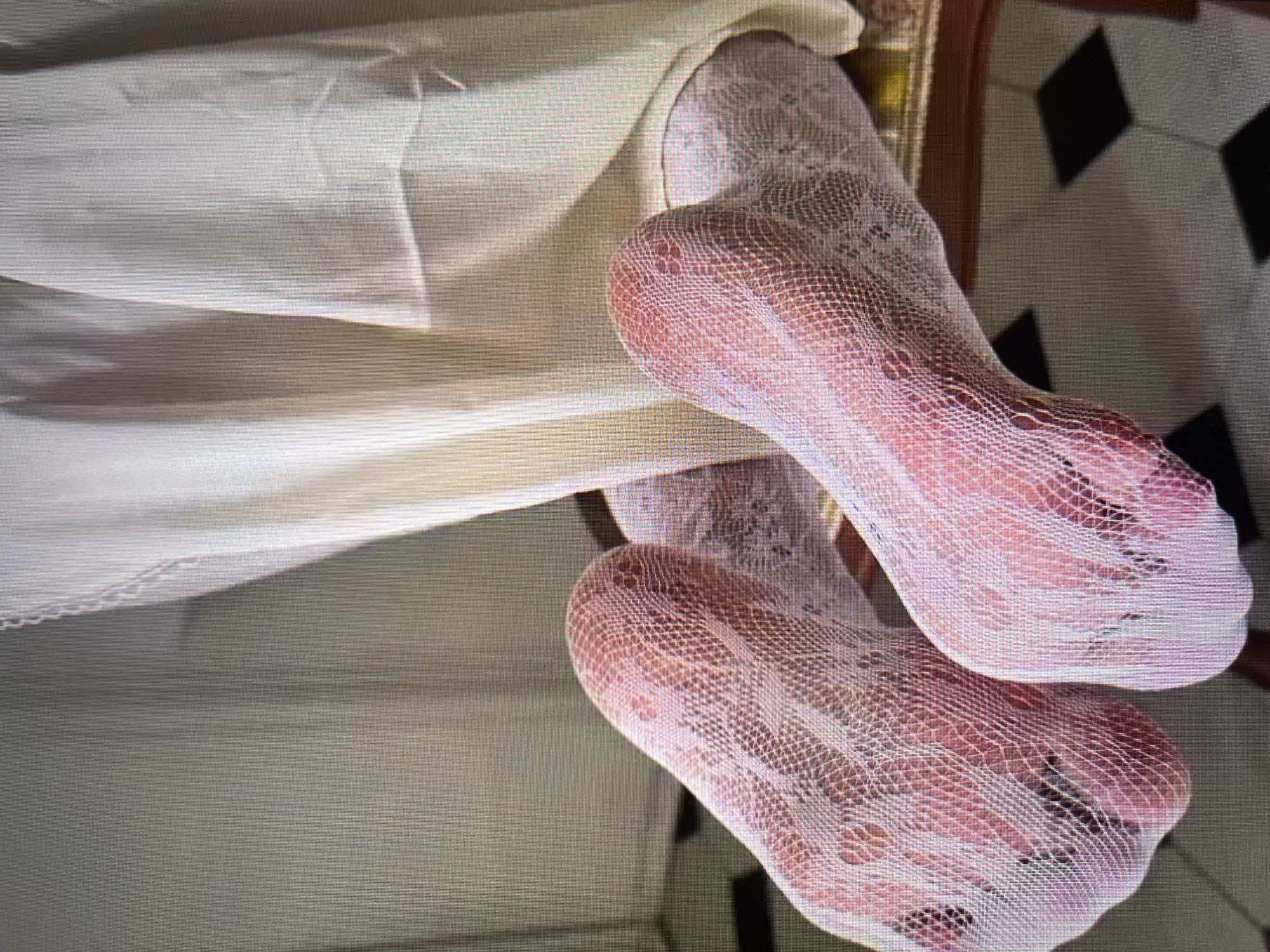 The image shows a close-up view of a person's feet wearing sheer, pink patterned stockings. The feet are resting on what appears to be a chair with a patterned floor visible in the background. The person is wearing a long, light-colored garment, likely a dress or skirt, that drapes over their legs and feet. The overall impression is one of indoor, domestic setting. There is no information in the image to suggest a location in Simferopol, Ukraine, or any specific city.