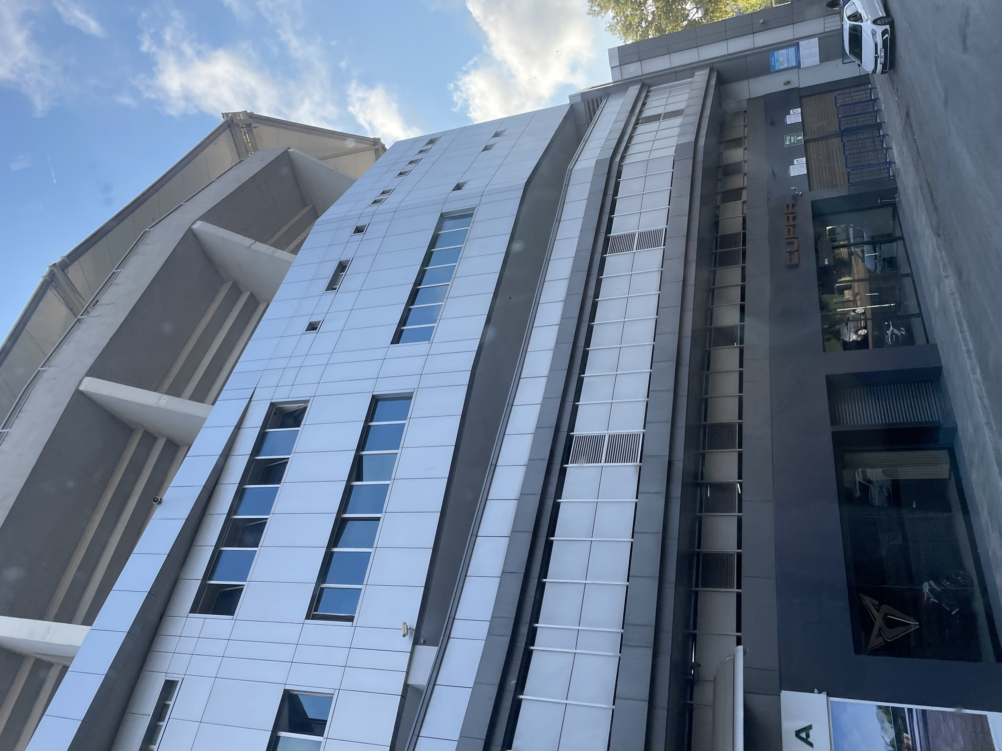 The image shows a modern building in Kadıköy, Turkey, with a distinctive curved facade. The upper levels of the building are clad in silver metal panels and feature numerous windows. Above this, the tiered seating of a stadium structure is visible, supported by concrete pillars. The lower levels of the building appear to be a car dealership, with large glass windows displaying cars inside and a prominent "CUPRA" logo. A white car is parked on the street in front of the building. The sky is blue with scattered clouds. The scene is captured from a low angle, looking upwards.