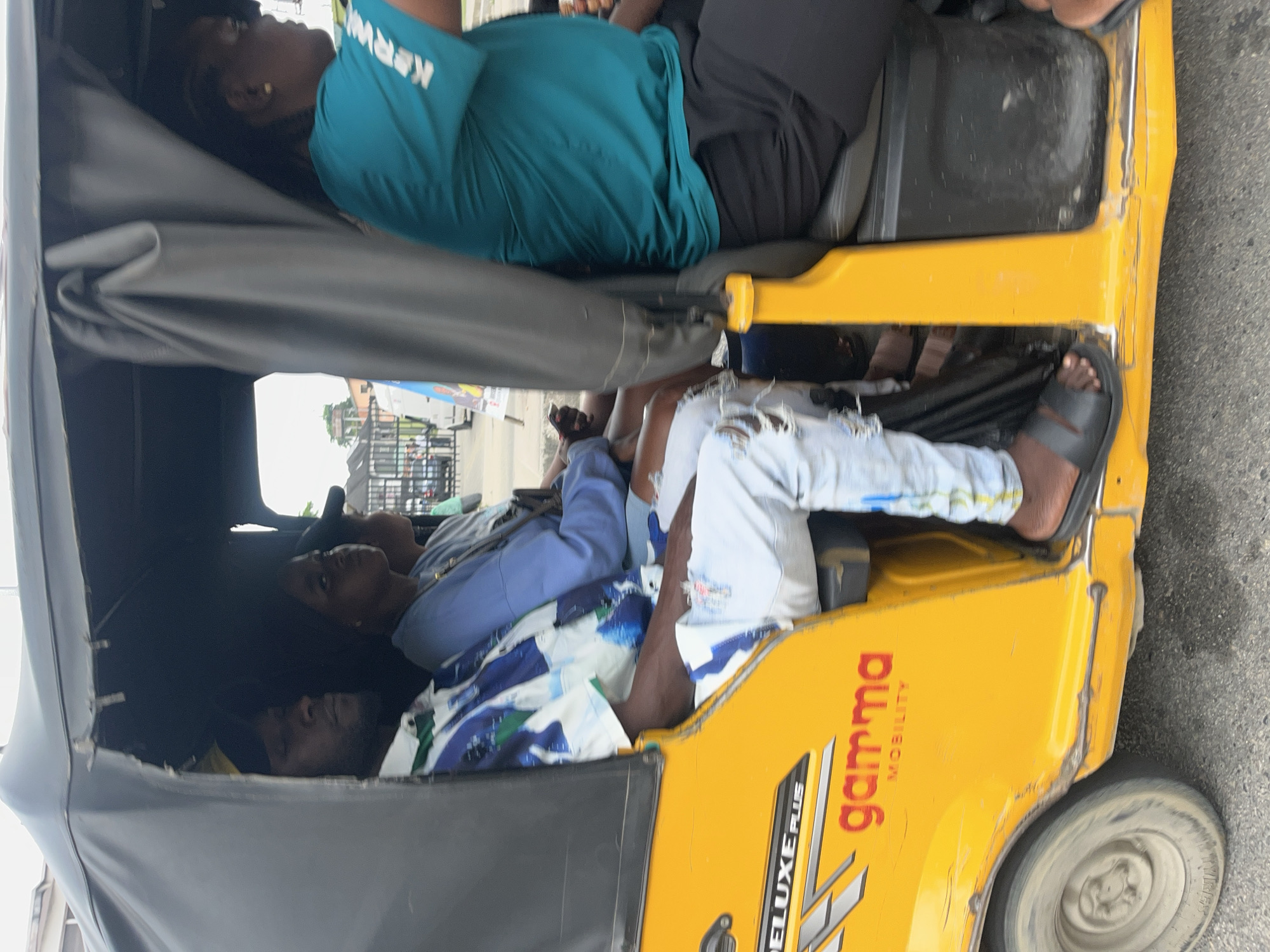 In Ado, Nigeria, a yellow motorized rickshaw, commonly known as an "okada" or "keke," transports four individuals. The rickshaw has a black canvas roof and a bright yellow exterior adorned with the text "DELUXE PLUS" and "GAMMA MOBILITY" in red lettering.
The occupants, viewed from the right side of the rickshaw, include a woman in the driver's seat wearing a teal t-shirt with white text that appears to read "KERWA." She is seated in the front passenger seat, which is likely the driver's position. Behind her, a young woman in a light blue hoodie and black pants sits on the left side of the passenger bench.
Next to her, a man wearing a blue, white, and green patterned short-sleeved shirt and distressed light blue jeans sits in the middle. He is looking forward, with his left arm resting on his lap. He is wearing black slides on his feet. The fourth passenger, a woman, is partially visible in the front passenger seat on the far right, wearing a teal shirt and black pants. Her left arm is resting on her lap.
The rickshaw is in motion, indicated by the blur of the road and the slight motion blur on the occupants. The background shows a street scene with buildings and other vehicles, suggesting an urban environment. The lighting suggests it is daytime, possibly overcast or late afternoon, given the soft shadows.