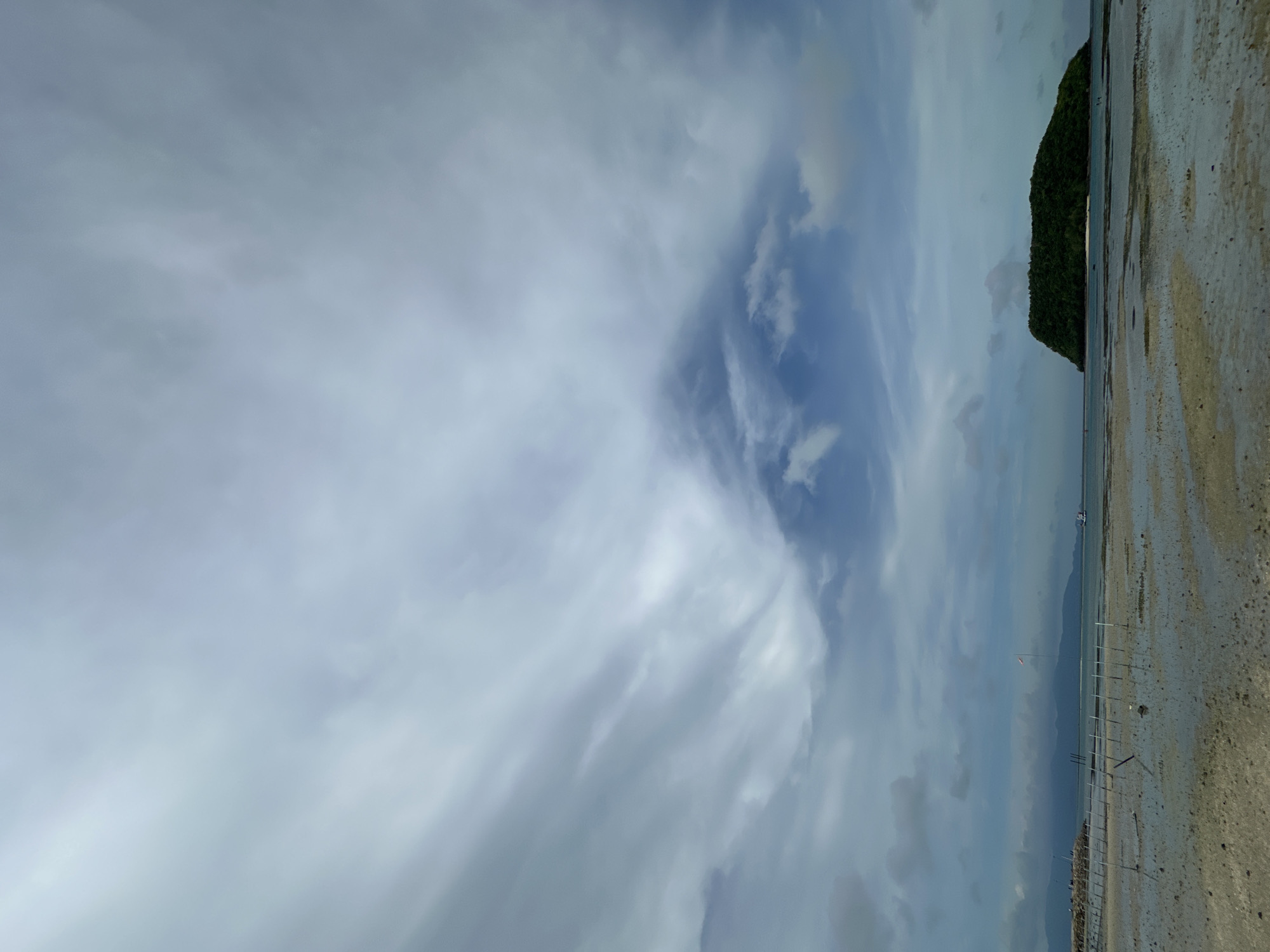 The image depicts a coastal scene on a cloudy day. In the foreground, a sandy beach is wet and reflects the sky, with shallow water covering parts of it. Several thin, vertical poles stick out of the sand, arranged in rows, possibly marking a boundary or a fishing area. In the mid-ground, the calm sea stretches towards the horizon. To the left, a breakwater or jetty extends into the water, with a few indistinct figures and structures visible on it. A small boat is seen in the distance. To the right, a lush green, uninhabited island with a rounded top rises from the sea, appearing as a dominant natural feature. The sky is filled with dramatic, striated clouds, predominantly light gray with some darker patches and hints of blue sky peeking through. The overall atmosphere is overcast and serene.