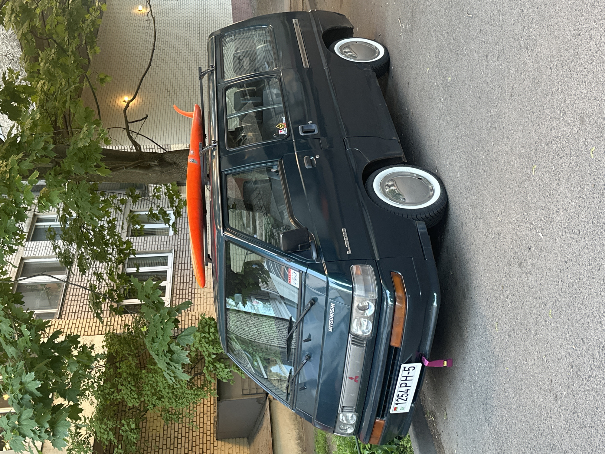 A dark-colored Mitsubishi L300 van is parked on a paved street in what appears to be a residential area. The van has a Belarusian license plate that reads "1254 PH-5." An orange surfboard is strapped to the roof rack. The van's tires have white sidewalls and chrome hubcaps. The background includes a multi-story brick building with windows and trees with green foliage. Based on the license plate, the location is likely Minsk, Belarus. No people are visible in the frame.