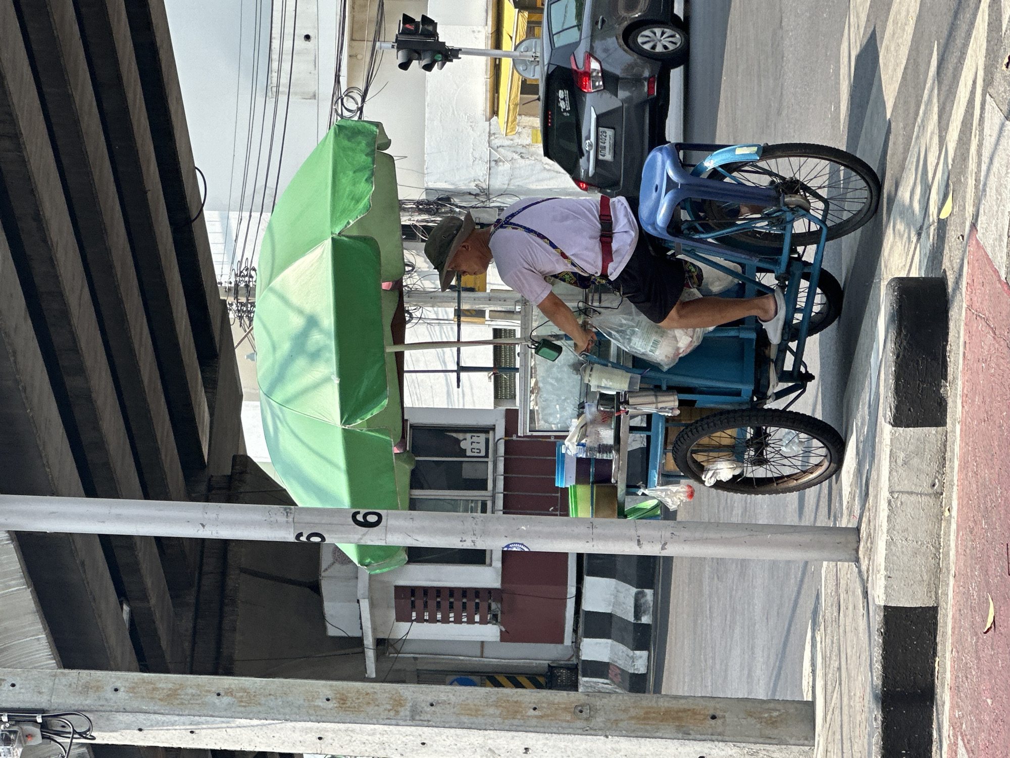 An older man operates a mobile street vendor cart under an elevated concrete structure in Bangkok, Thailand. The man, seen from the side, wears a wide-brimmed hat, a light-colored t-shirt with colorful patterned suspenders, dark shorts, and white sneakers. He is seated on a blue plastic chair attached to his tricycle-style cart.
The vendor cart is equipped with a large, bright green market umbrella providing shade. Various containers, plastic bags, and items for sale are visible on the cart. In the background, a dark grey car with a Thai license plate ("4กณ 4029") is parked on the street. A traffic light is visible further down the road.
The setting is an urban environment, likely under a bridge or elevated railway. The ground features an asphalt road, a sidewalk with black and white striped curbing, and a red-painted section. A prominent grey utility pole with the number "6" (or "9") partially visible runs vertically through the frame. Overhead, numerous electrical wires crisscross against the bright sky. The lighting indicates a sunny day, casting strong shadows on the ground and structures. Buildings with windows and a mix of concrete and darker panels are visible behind the vendor.