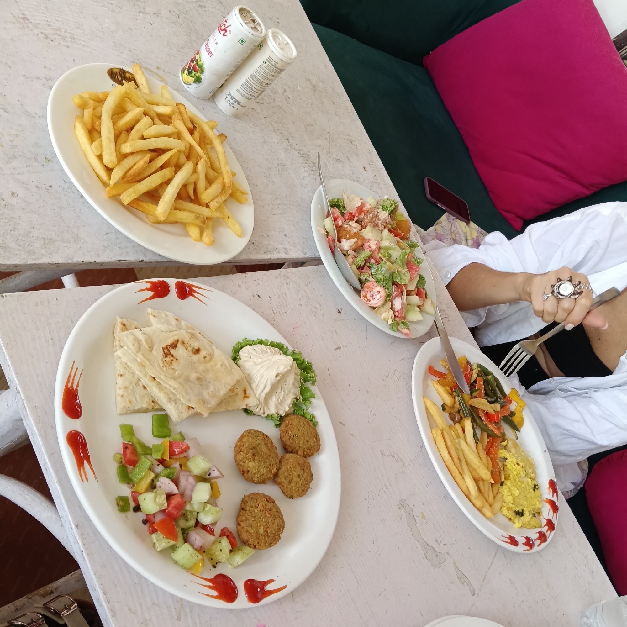 A table in Mandrem, India, displays three plates of food and two containers. The first plate has french fries. The second plate contains a salad of lettuce, tomato, and cucumber. A woman in a white shirt and dark pants is eating from the third plate with a fork and knife. The plate contains a scramble-like mixture, french fries, and mixed stir-fried vegetables. A plate containing pita bread, hummus, falafel, and a fresh diced vegetable salad sits adjacent. Ketchup decorates both of these plates. Behind the woman is a green couch with a red pillow. A mobile phone is on the couch. Two canisters rest between the french fries and the salad. The table is white with a painted finish.