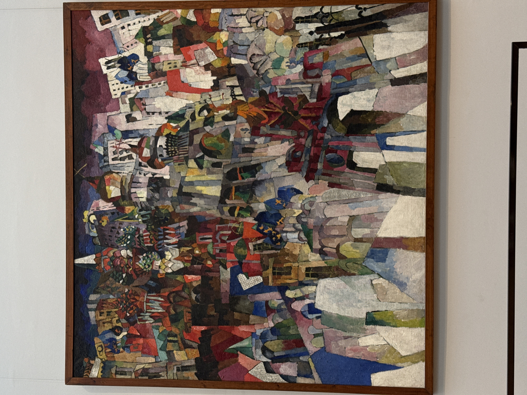 The image shows a framed abstract painting hanging on a white wall. The painting features a complex composition of geometric shapes and layered colors, primarily in shades of red, blue, brown, and white. The shapes are arranged in a dense, seemingly haphazard manner. The painting is surrounded by a simple, wooden frame. The scene is indoors, with a second darker frame partially visible on the right of the painting. Moscow, Russia cannot be determined from the image. There are no people visible.