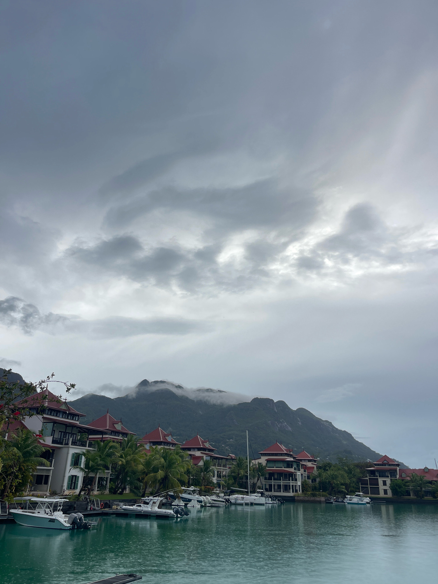 The image shows a waterfront view of apartments with red-tiled roofs and balconies, nestled amongst palm trees. Several boats are docked in the turquoise water in front of the buildings. In the background, a large, mist-shrouded mountain range rises above the scene, under a cloudy sky. The location is identified as Providence, Seychelles.