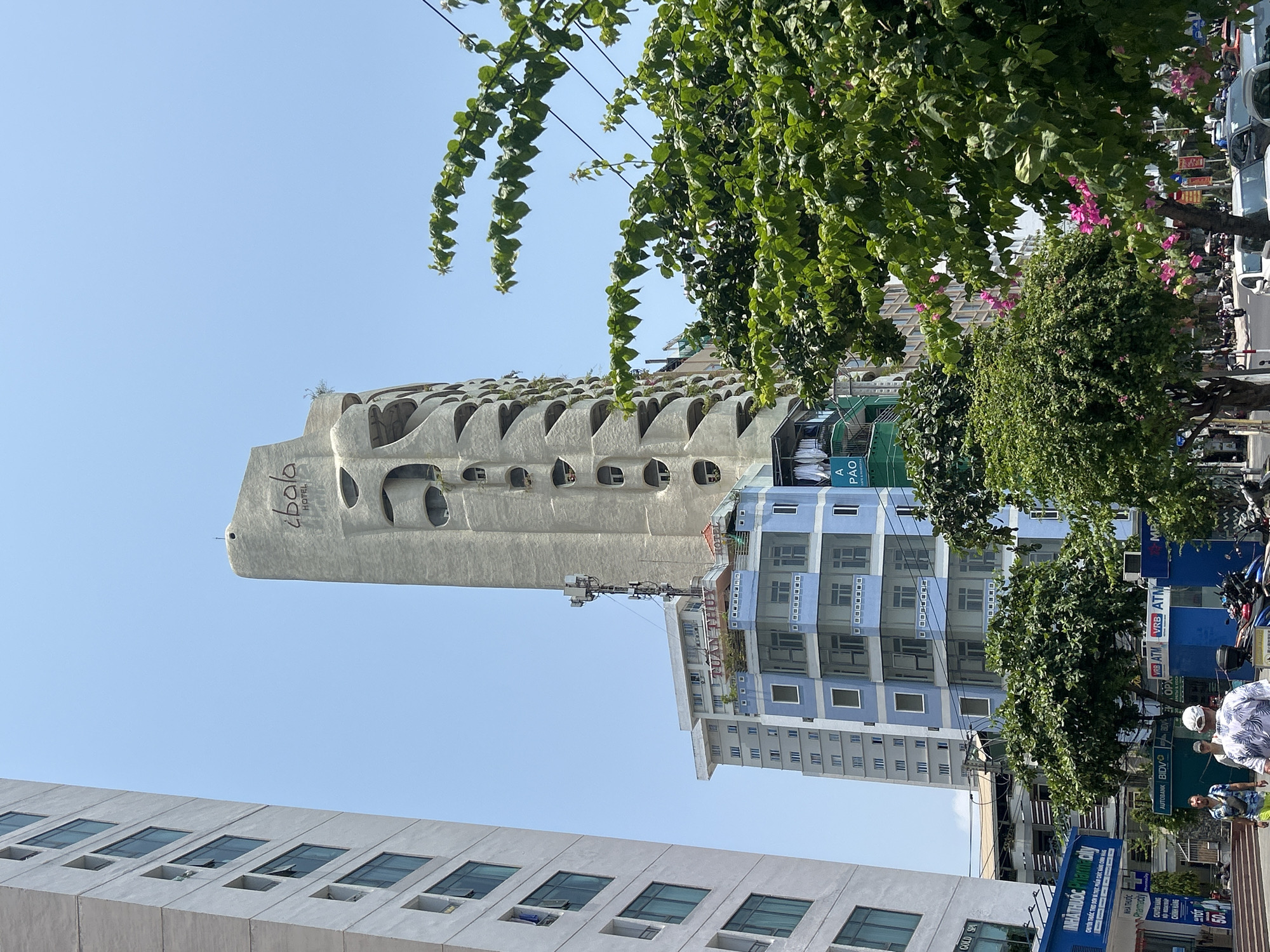 The image depicts a street scene in Bắc Nha Trang, Vietnam. The architectural landscape is dominated by several buildings: a unique, elongated structure with circular cutouts, identified as "ibola HOTEL" via signage; a blue and white apartment building; and a lower-rise building with a grid of windows. Green trees and foliage partially obscure the buildings. Street-level details include signage for a pharmacy and bank ATMs (VRB and BIDV). Pedestrians are visible on the right side of the image.