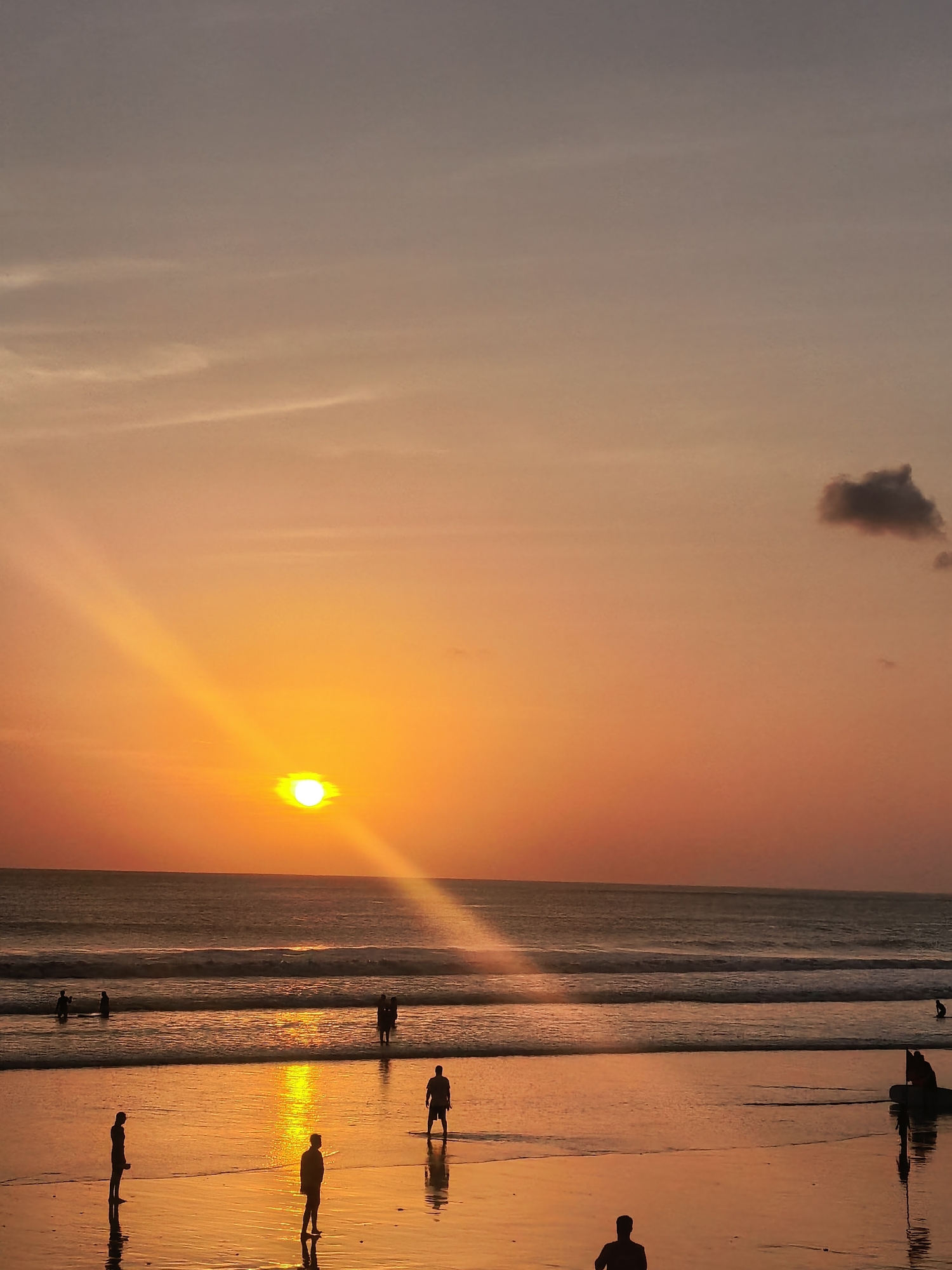 The image depicts a serene sunset on Kuta Beach, Bali, Indonesia. The sun, a bright orb, hangs low in the sky, casting a warm, golden glow across the scene. Its rays stretch across the water, creating a shimmering path. The sky transitions from a soft orange near the horizon to a pale, dusky purple higher up. A solitary dark cloud is visible in the upper right quadrant.
The ocean is calm, with gentle waves rolling onto the shore. Several people are scattered across the beach and in the shallow water. In the foreground, two figures stand on the wet sand, their reflections mirrored in the shallow water. Further back, near the water's edge, more individuals are visible, some wading, others standing. In the distance, more silhouettes can be seen near the surf. The overall atmosphere is tranquil, with the natural beauty of the sunset dominating the scene.